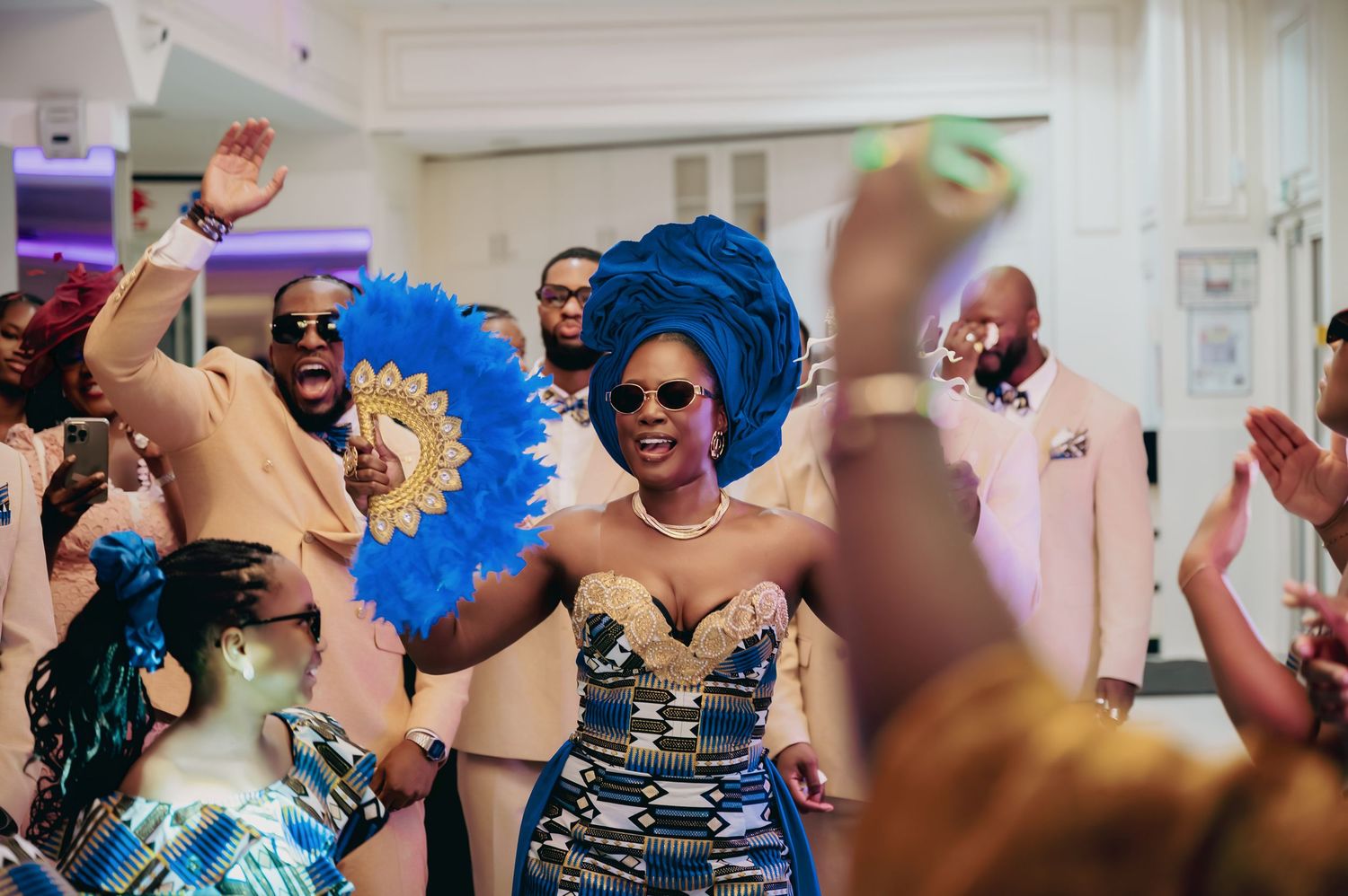 Bride in kente print dress dances joyfully with blue feather fan at African wedding reception.