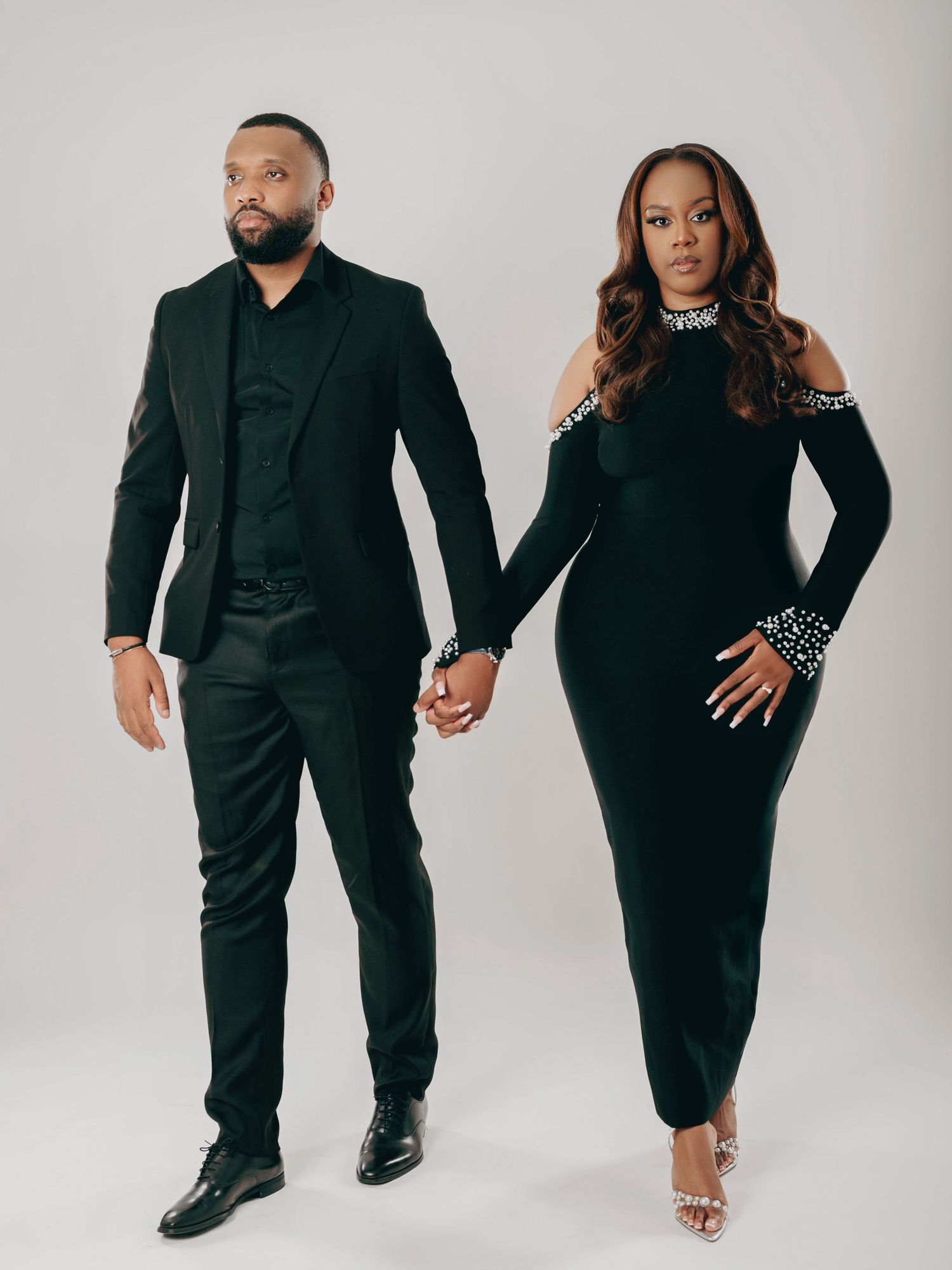 Couple in coordinating black formal outfits hold hands while posing in a professional studio.