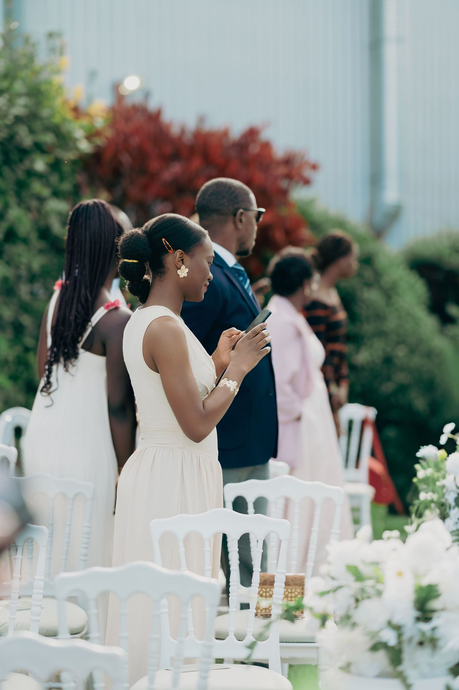 Wedding guests in formal attire standing among white chairs at an elegant outdoor garden ceremony.