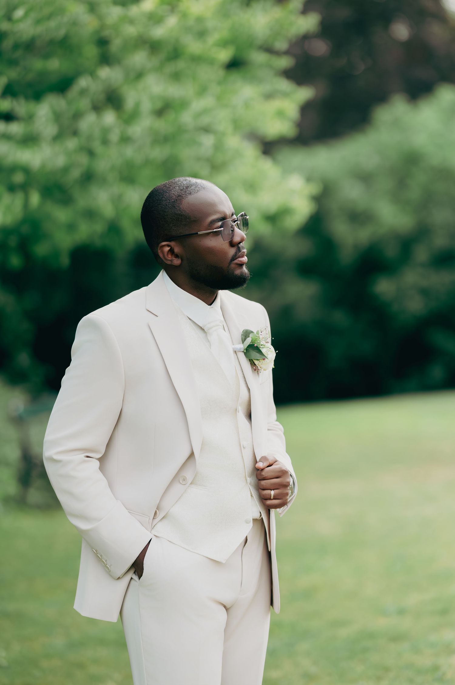 Stylish Black groom in a cream three-piece suit with boutonniere stands outdoors in lush greenery.