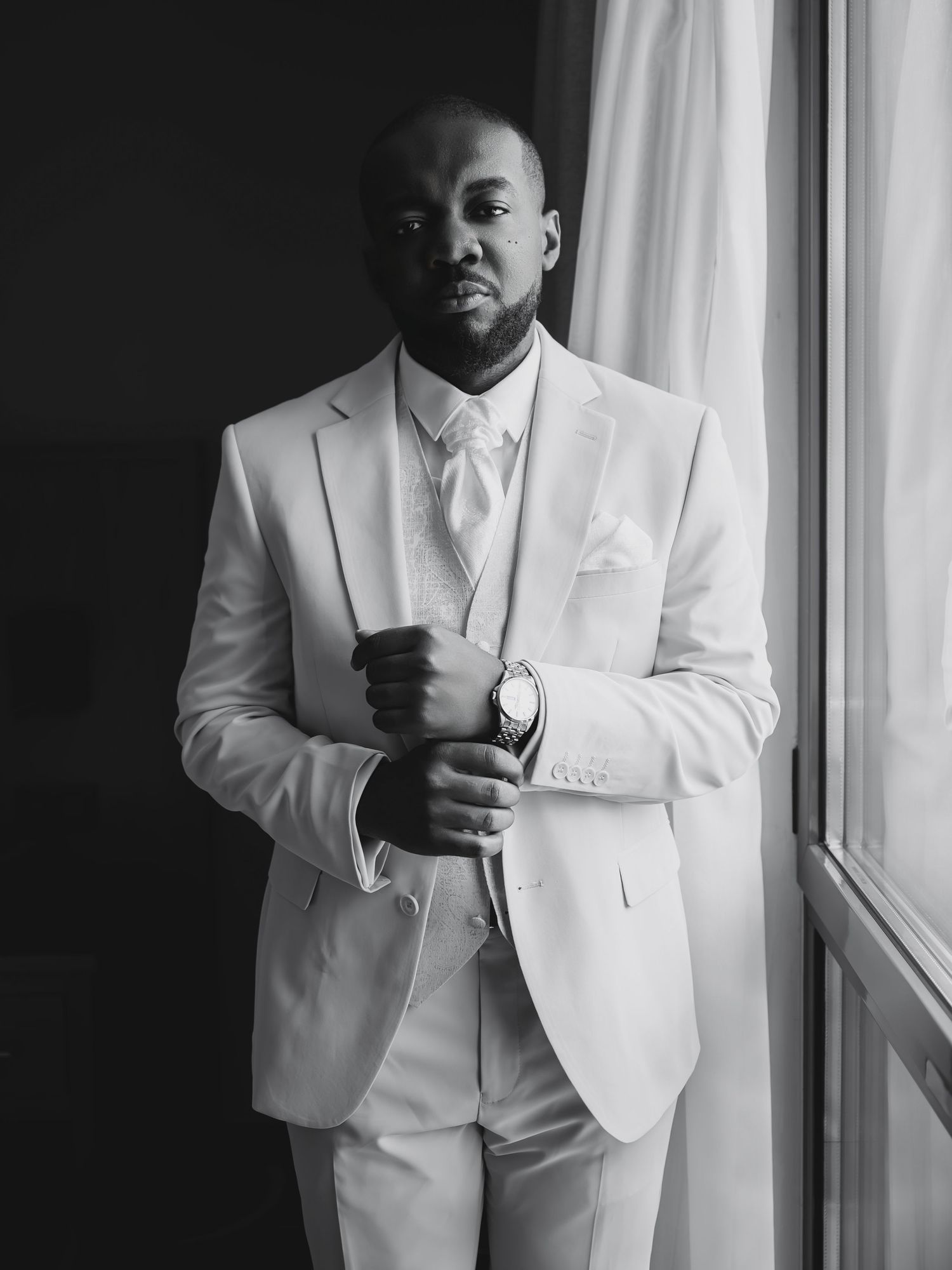 Groom in a white suit adjusting cufflinks in a black and white portrait.