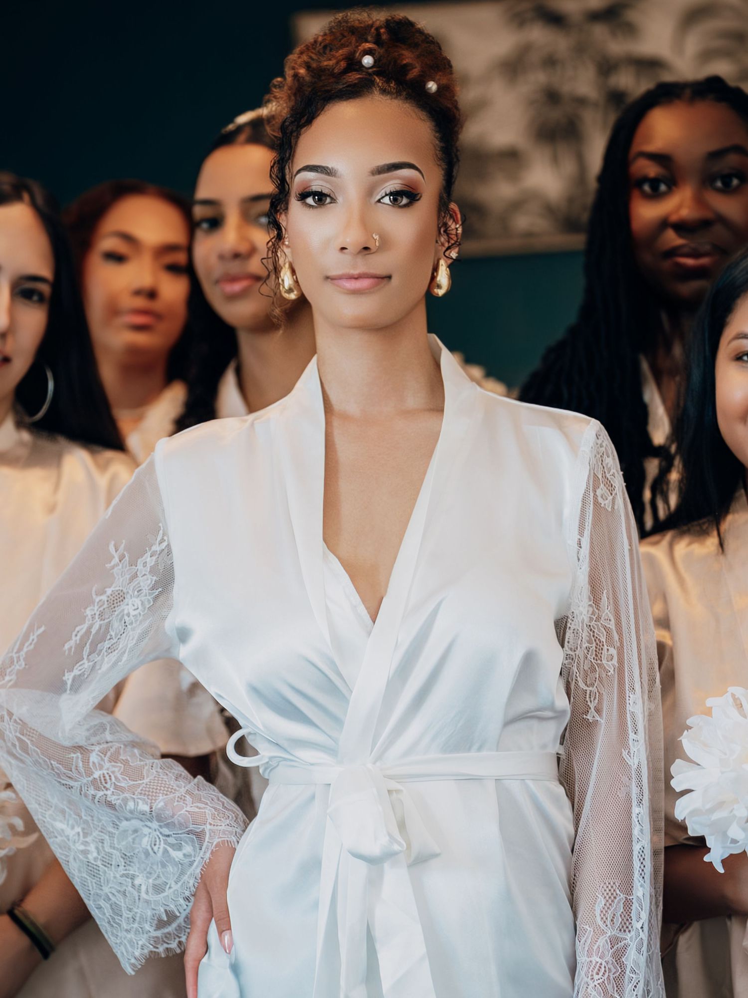 Stunning bride in white lace robe with pearl hair accessories stands before her bridesmaids in white robes.