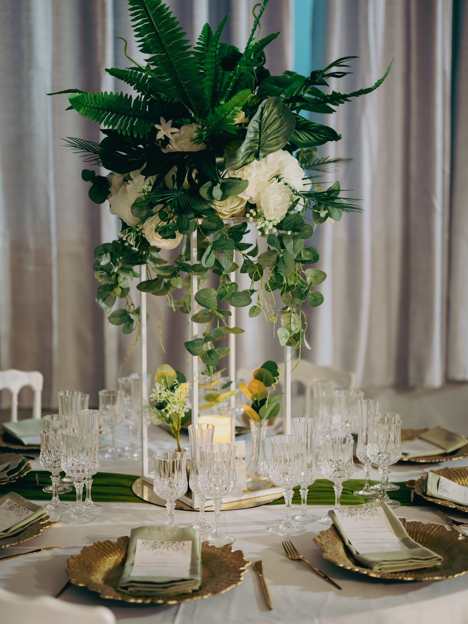 An elegant wedding reception table with tall greenery centerpiece, crystal glassware, and gold charger plates.