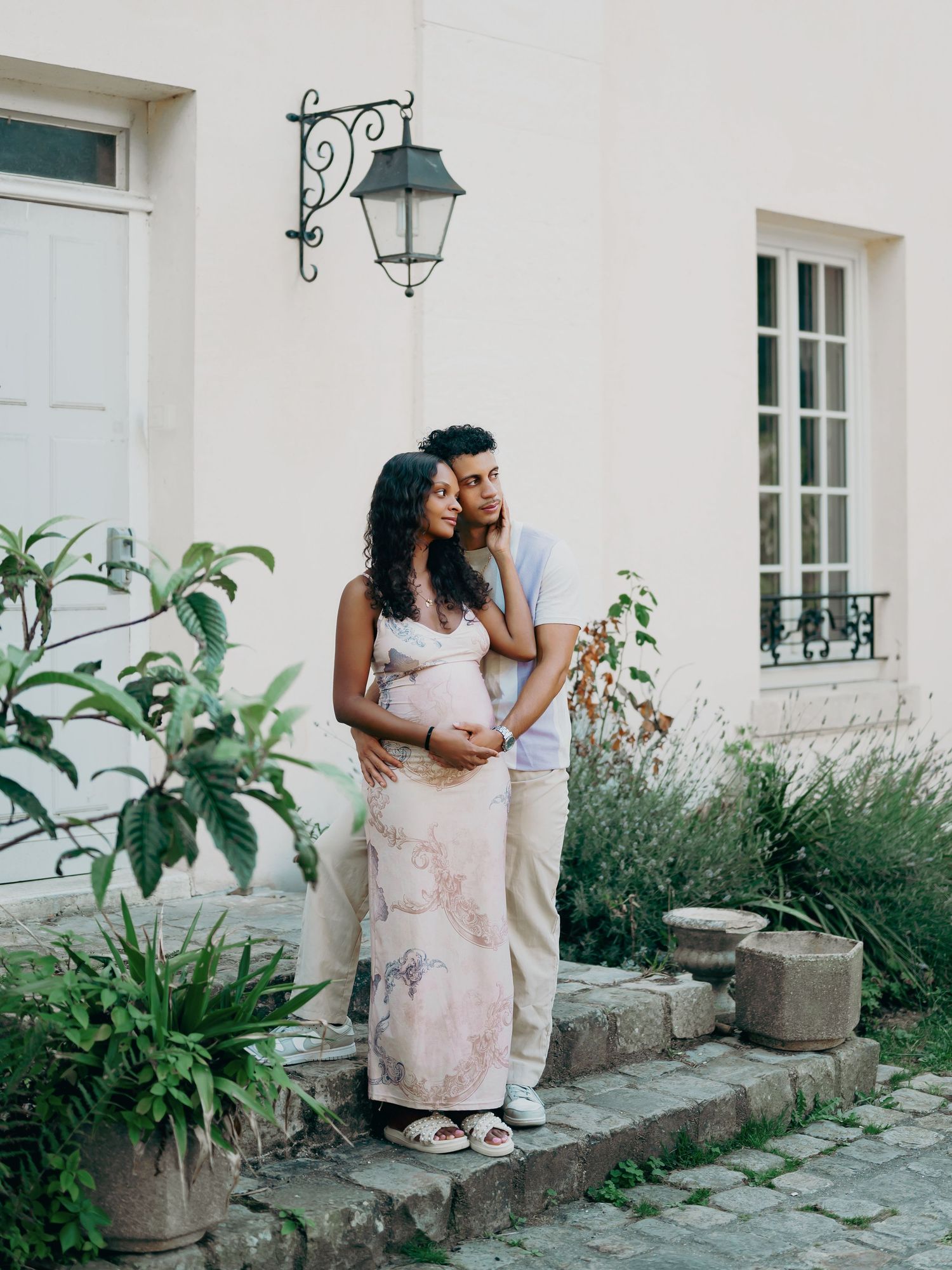 Pregnant couple embracing on stone steps outside a charming white French-style building with garden.
