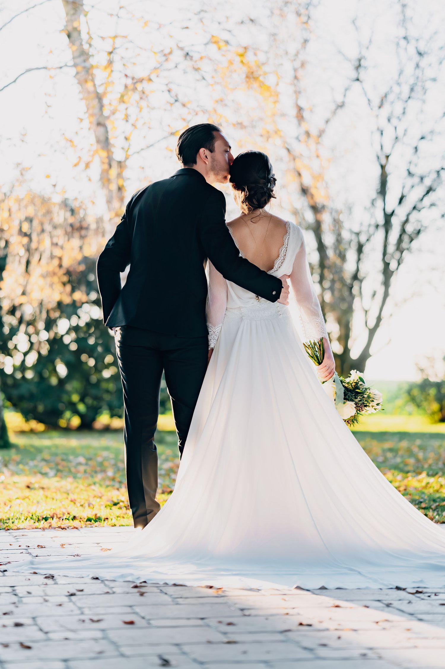 Groom kisses bride on cheek as they walk through a sunlit autumn park, her gown trailing behind.