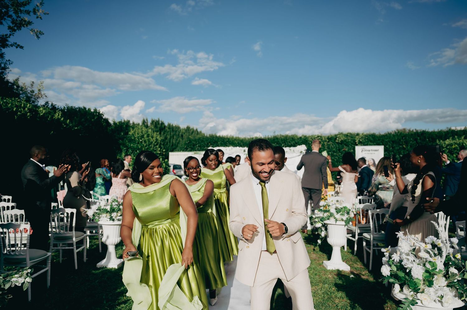 Groom in white suit celebrates with bridesmaids in green dresses at outdoor wedding ceremony.