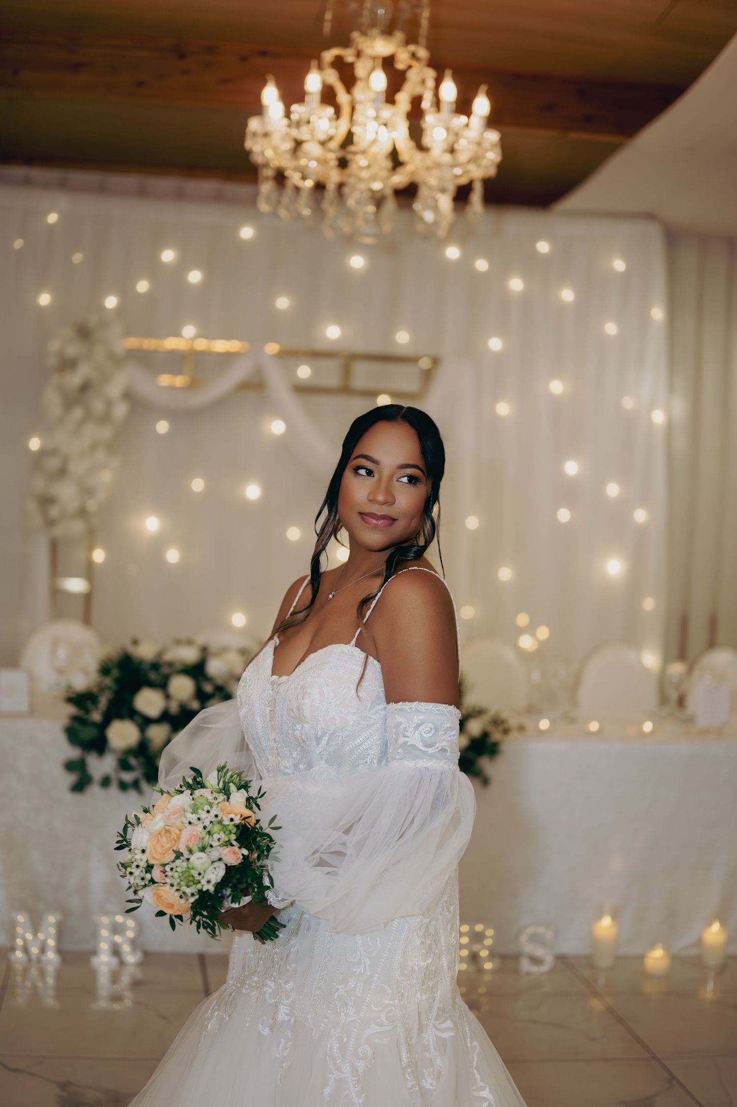 Elegant bride in lace wedding gown holding peach bouquet under crystal chandelier with twinkling fairy light backdrop.