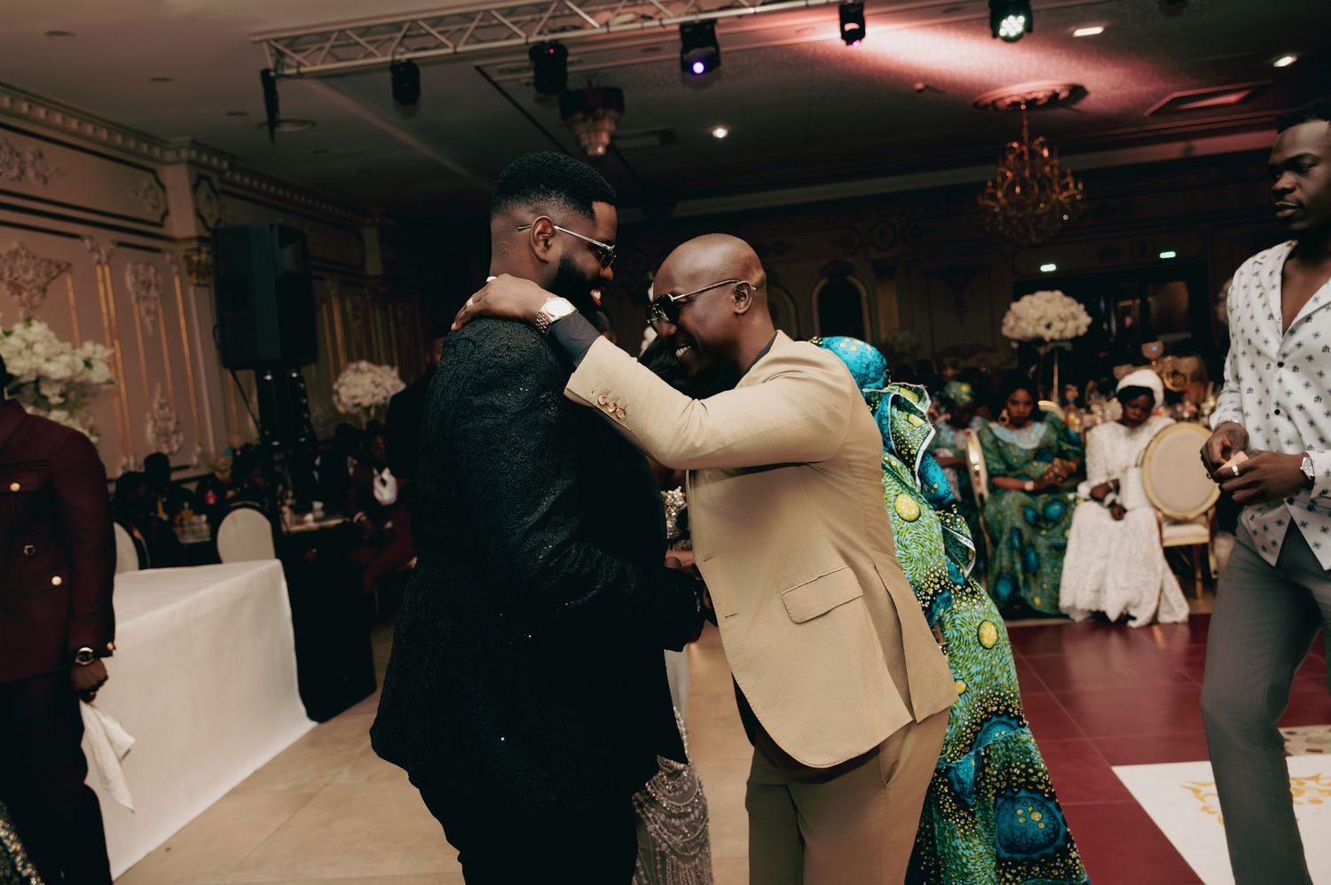 Groom embraces guest in tan suit during joyful wedding reception dance.