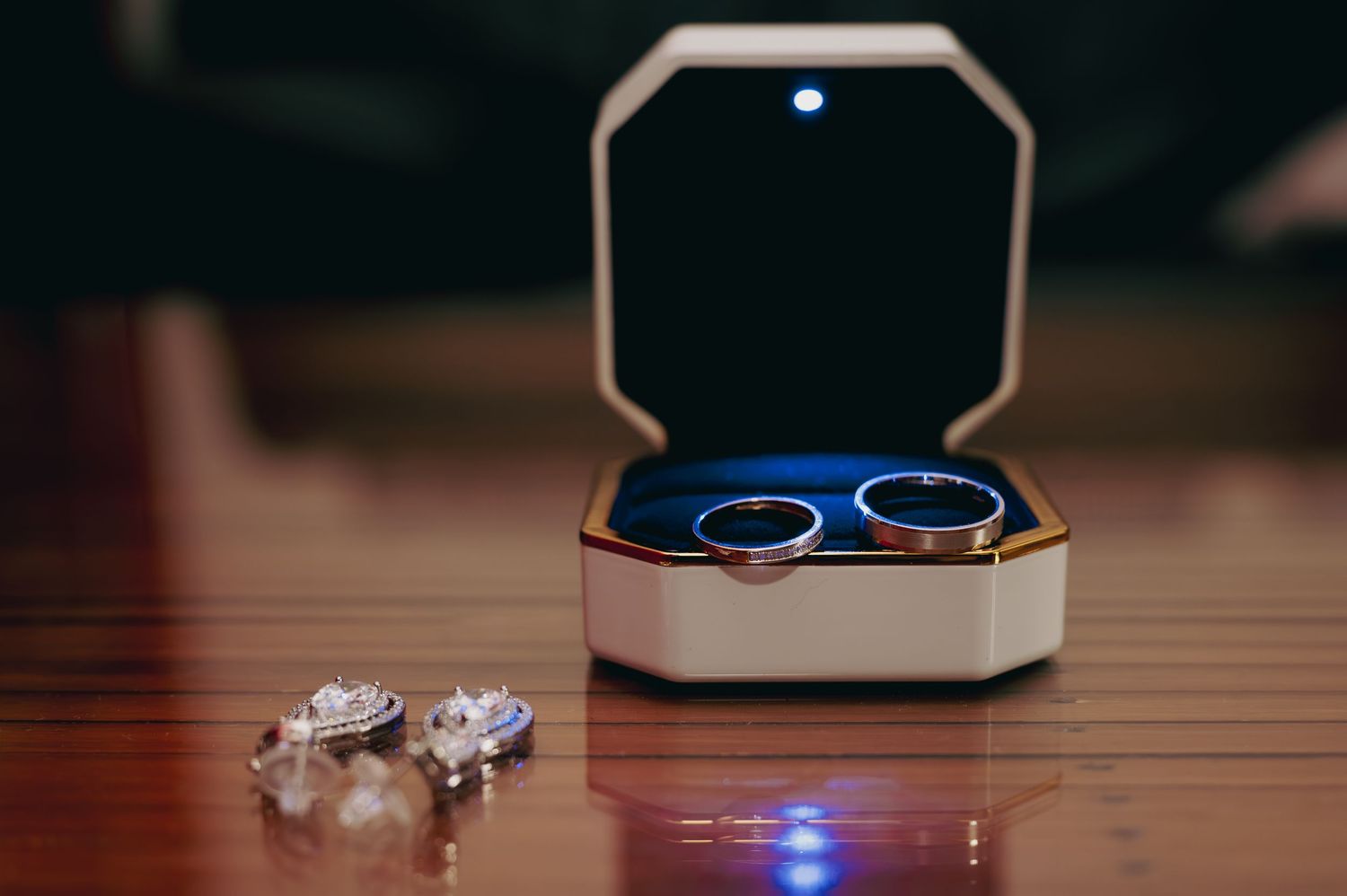 Two gold wedding bands displayed in a lit jewelry box with diamond earrings on wooden surface.