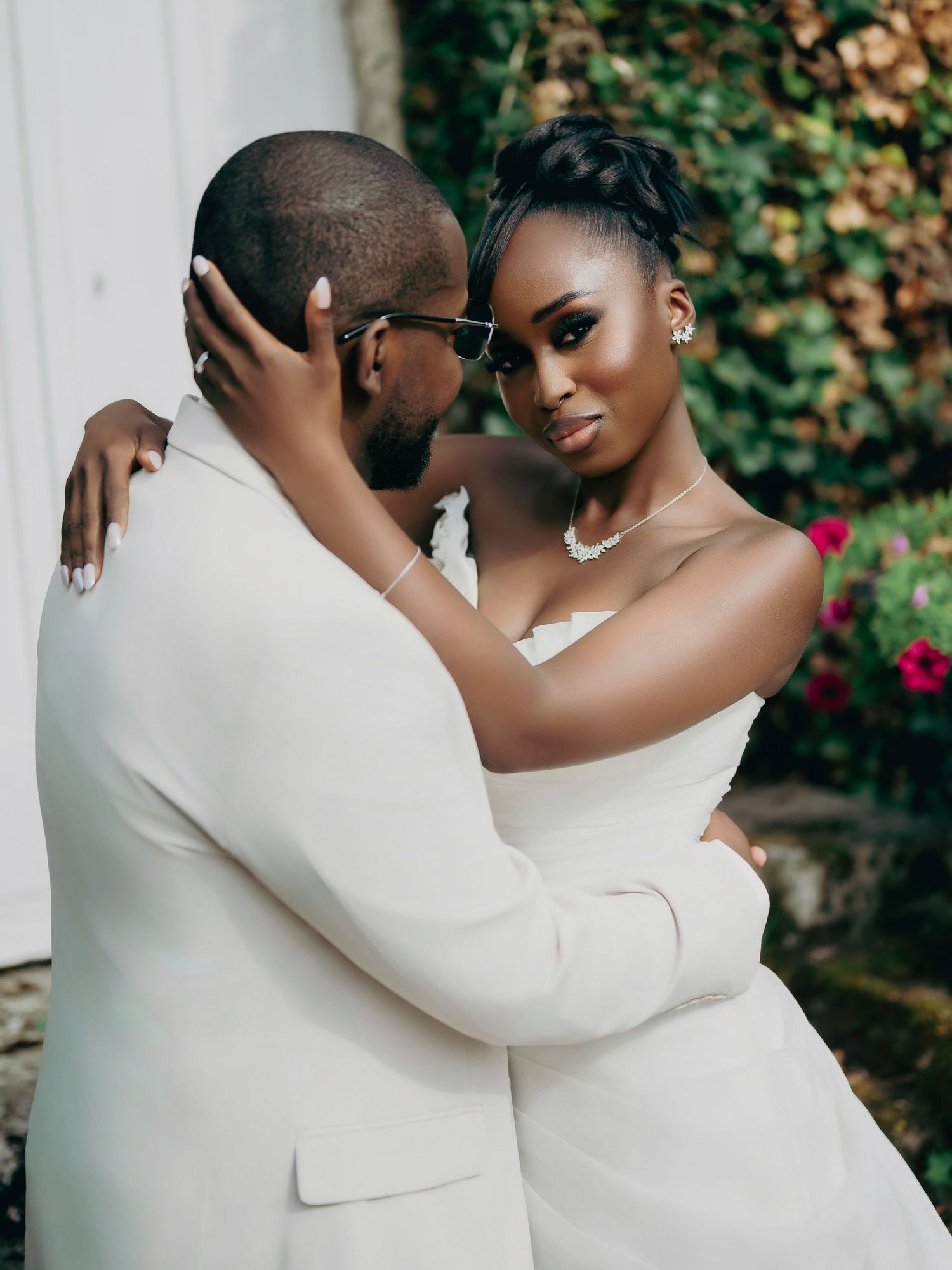 Bride and groom embrace intimately, bride gazing at camera wearing strapless gown and diamond necklace.