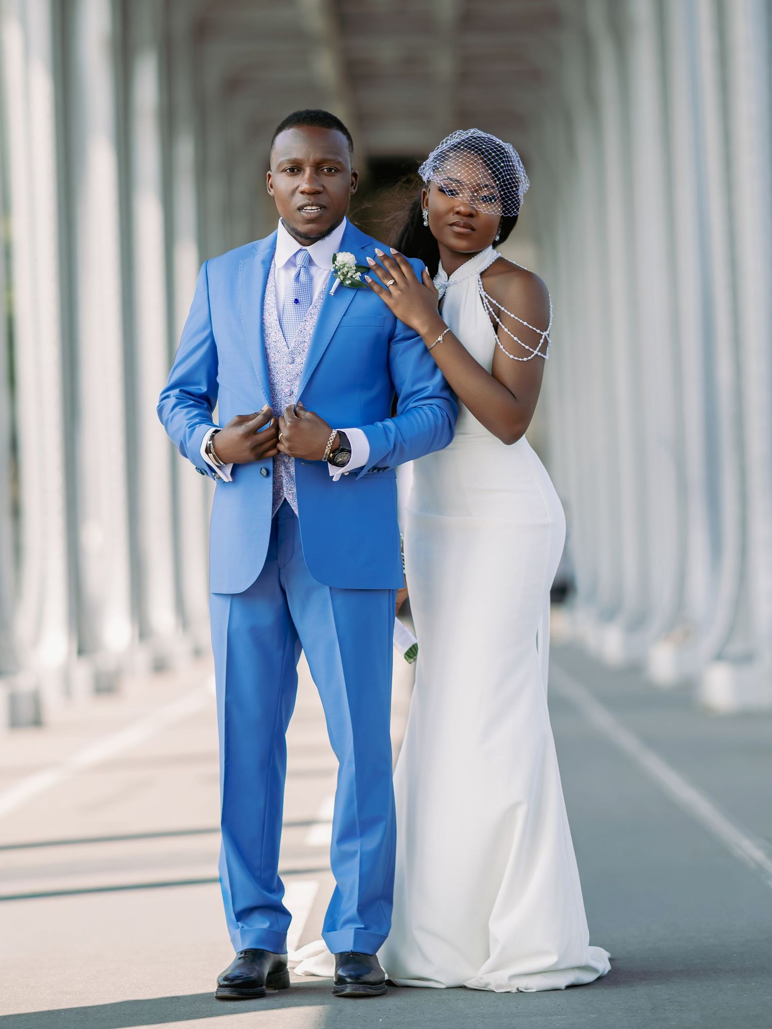 Black wedding couple poses in a white colonnade, groom in blue suit and bride in sleek white gown with birdcage veil.