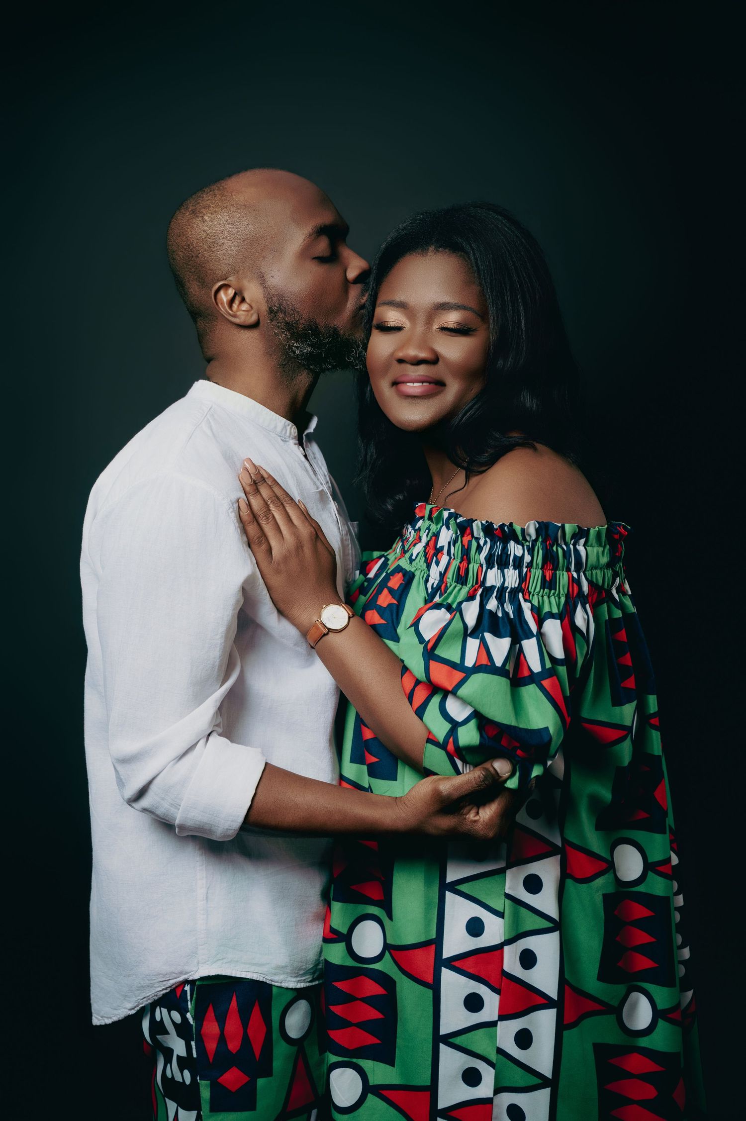 A man tenderly kissing a woman on the cheek as she smiles, both in African-inspired outfits.