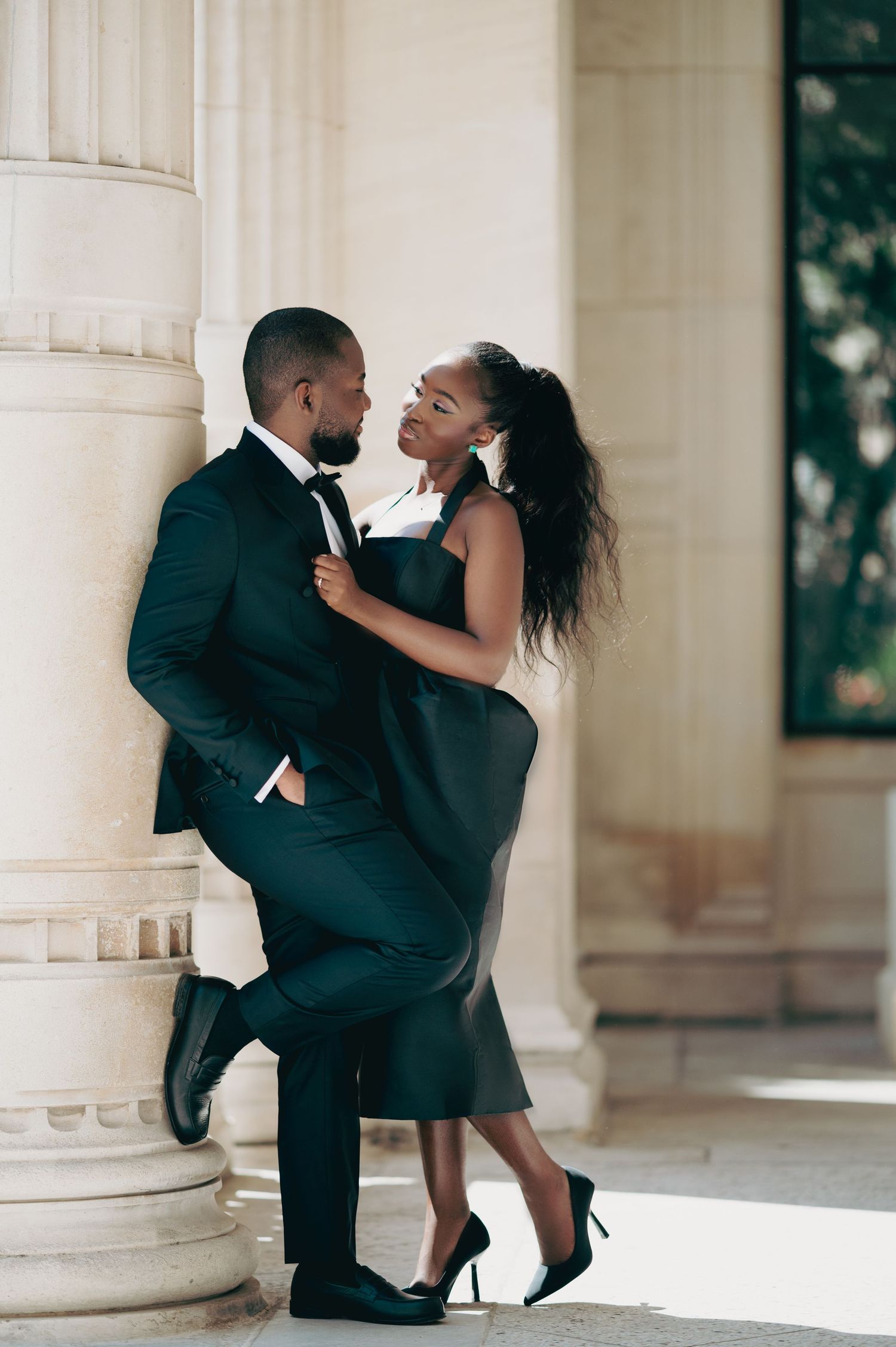 Stylish Black couple in black formal attire posing romantically between classical stone pillars outdoors.