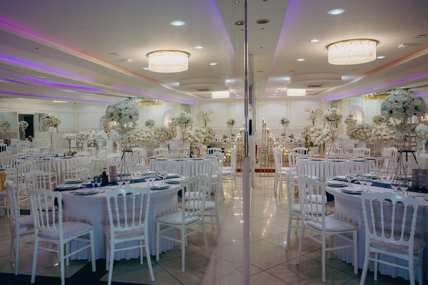 Elegant white wedding reception hall with Chiavari chairs, floral centerpieces, and soft purple lighting.