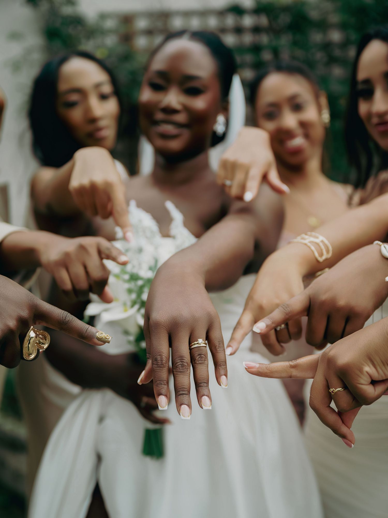 Joyful bride and bridesmaids in white pointing to wedding rings in fun group photo outdoors.