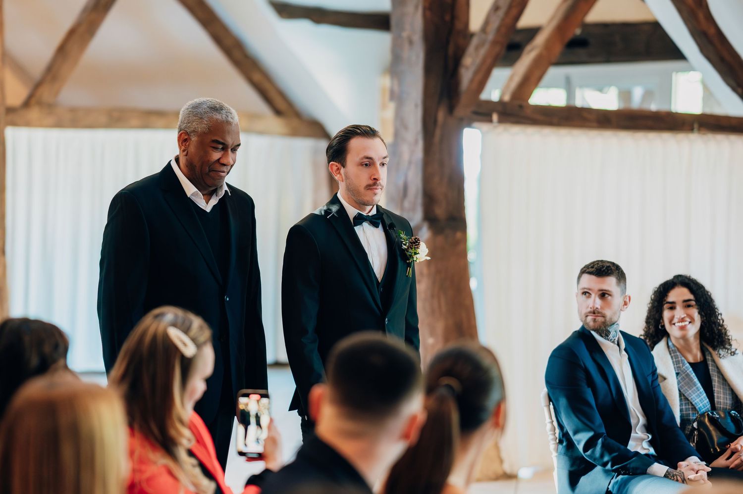 Groom in black tuxedo standing at the altar with guests seated in white chairs during a rustic barn wedding ceremony.