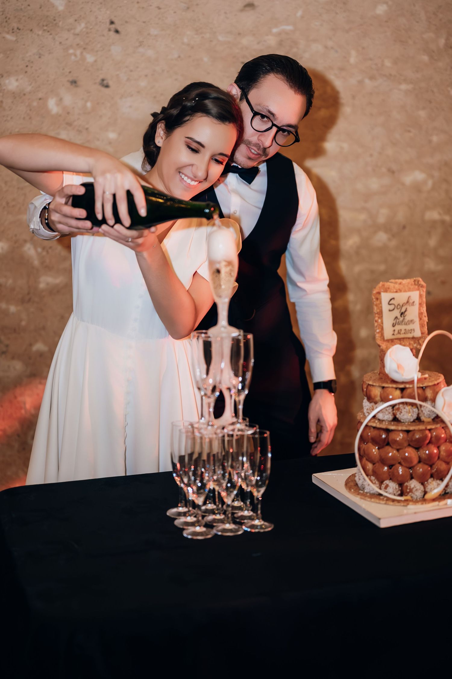 Bride and groom pouring champagne tower at wedding reception with croquembouche cake nearby.