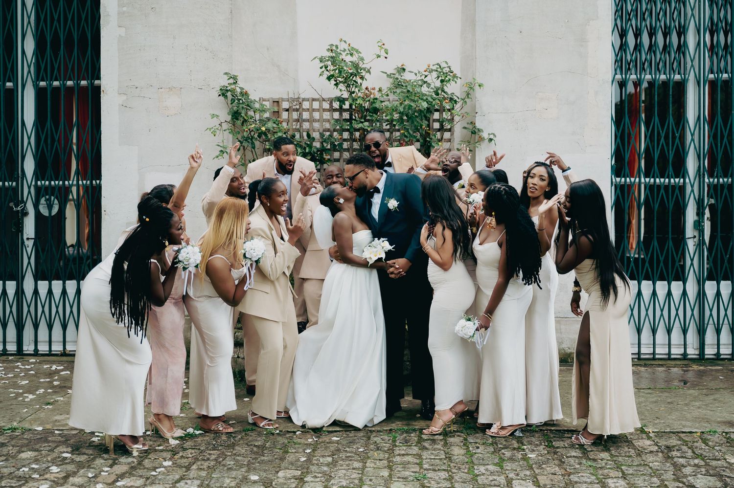 Newlyweds kissing surrounded by cheering bridesmaids and groomsmen on cobblestone courtyard.