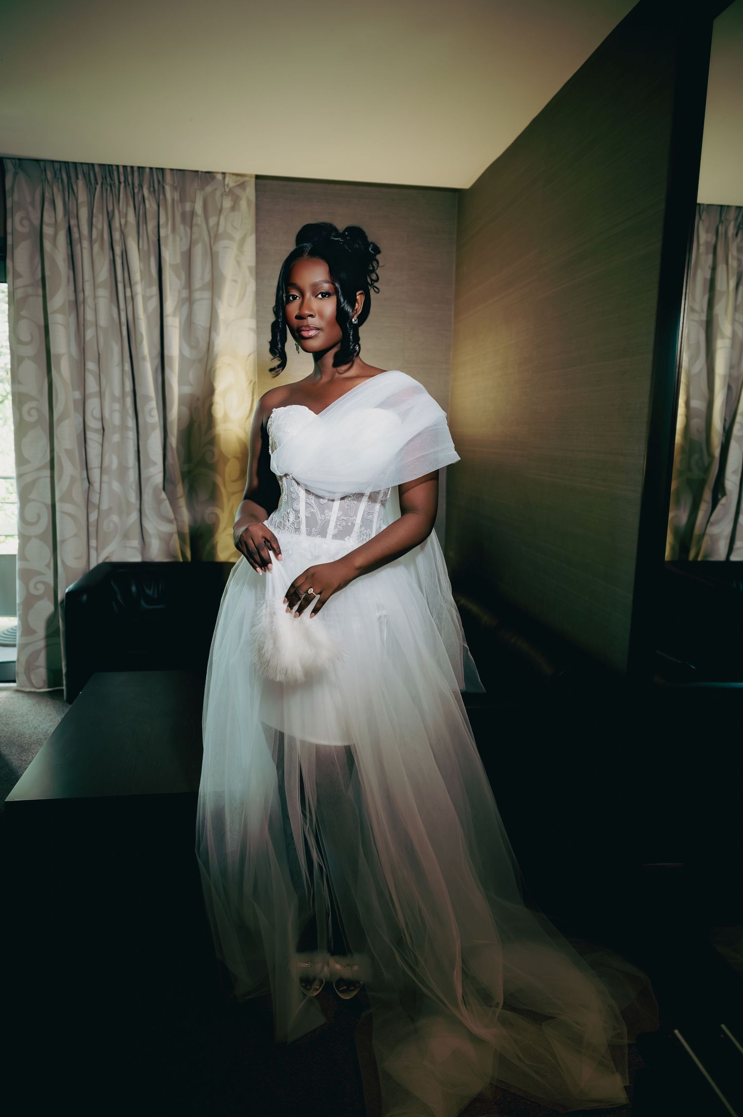 Elegant Black bride in white one-shoulder tulle corset wedding dress posing in hotel room.