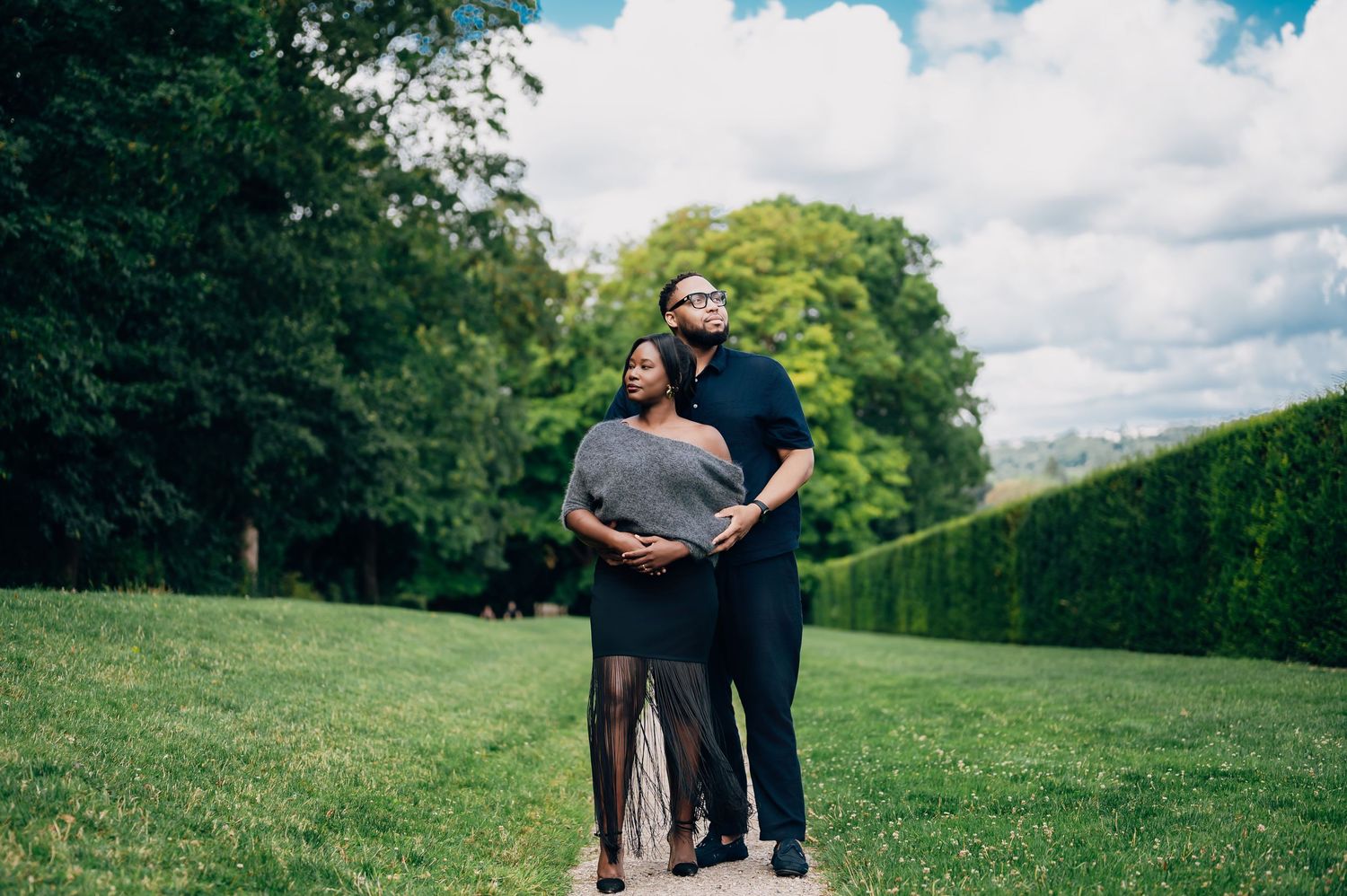 Couple standing close together on garden path, both looking up at cloudy sky with green hedges behind.
