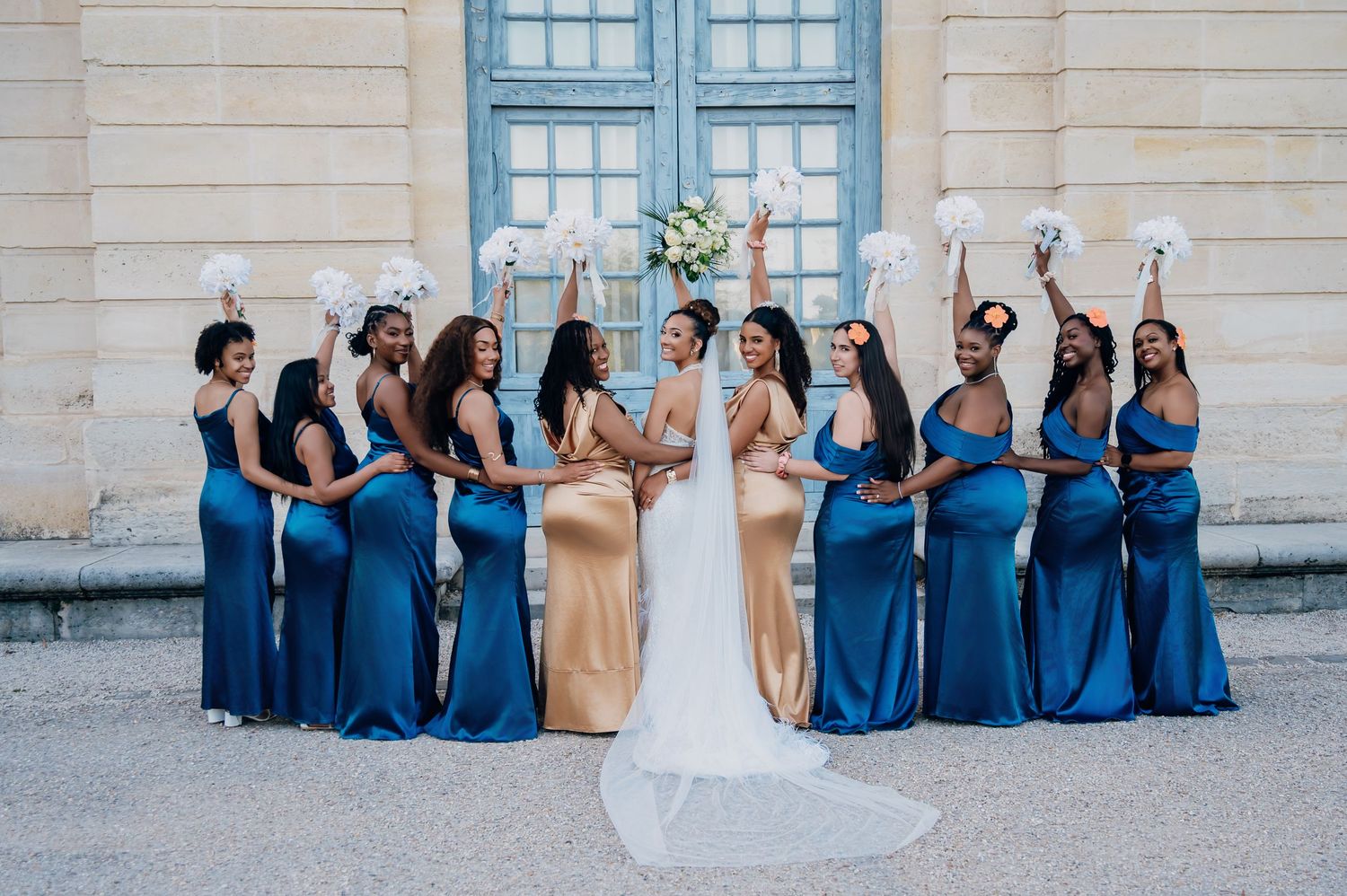Bridesmaids in blue and gold satin gowns cheer with white bouquets behind the bride.