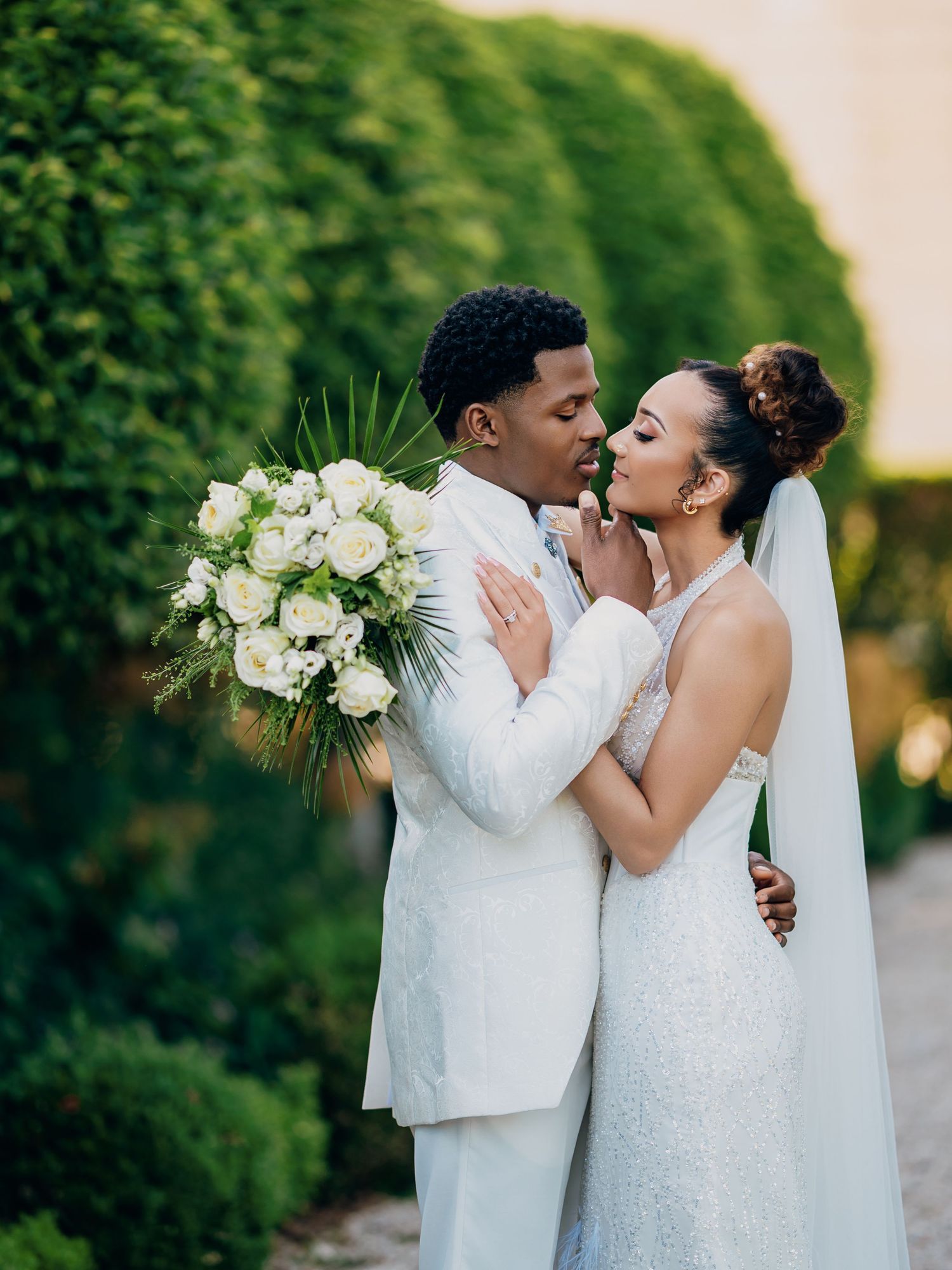 Couple shares intimate moment in garden, bride in lace gown with veil and groom in white suit with bouquet.