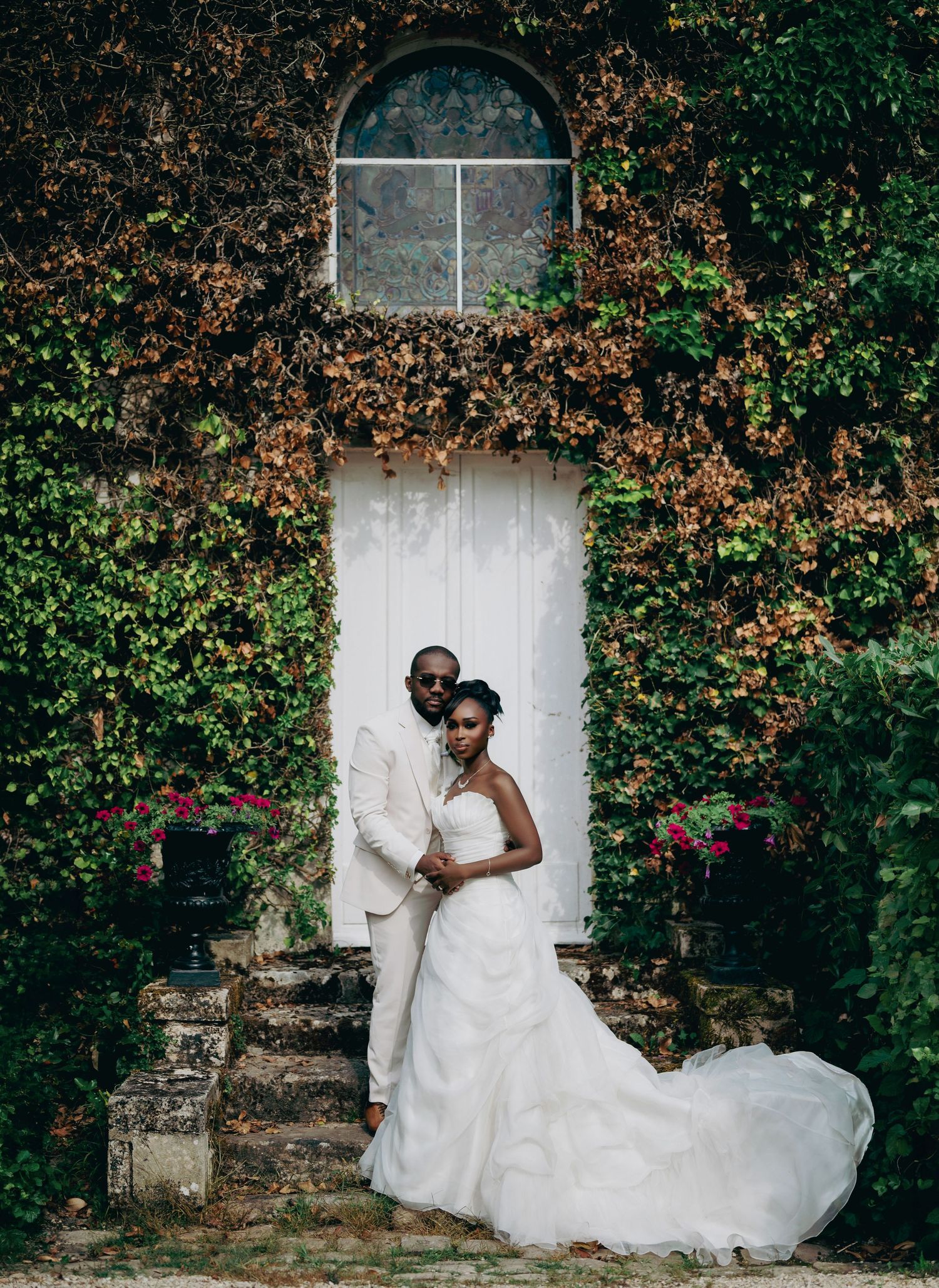 Bride and groom embracing before ivy-covered stone building with white door in garden.