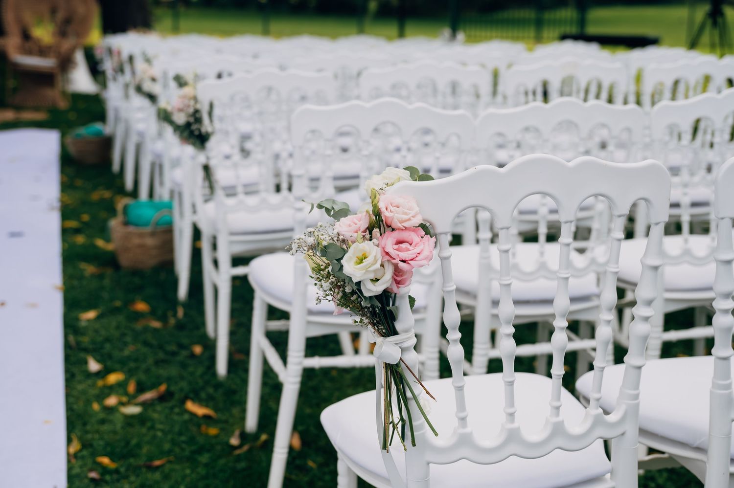 Outdoor wedding ceremony setup with elegant white chairs and a pink and white floral bouquet tied to the aisle seat.