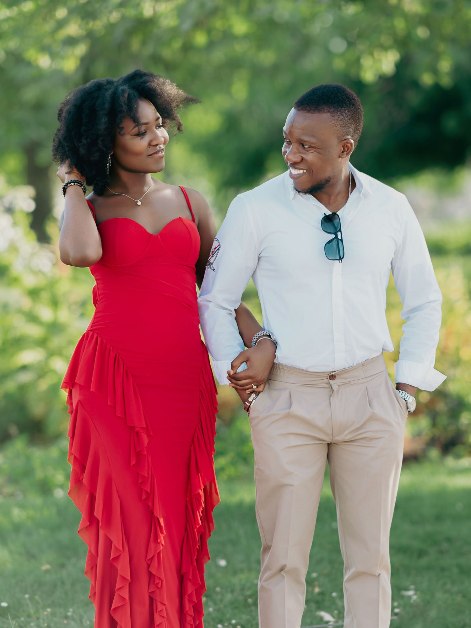 Black couple walking hand in hand through a sunlit green park, woman in red gown and man in white shirt.