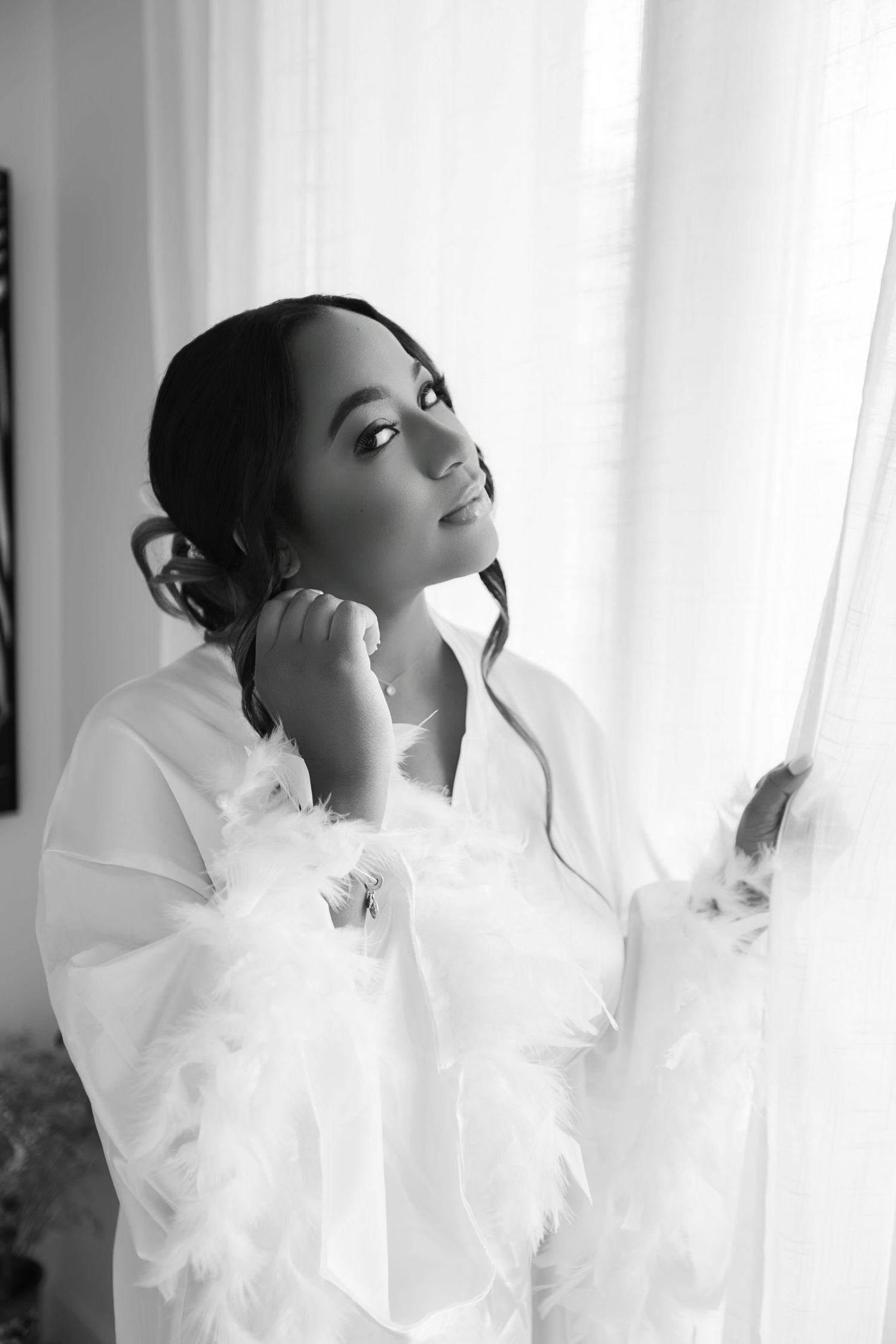 Black and white portrait of Black bride in feathered white robe gazing upward by sheer curtains.
