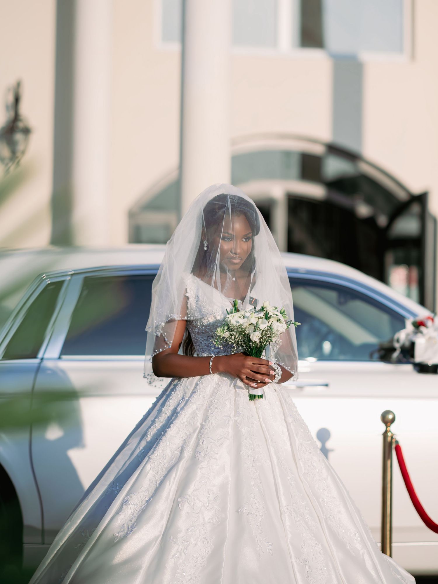 Elegant bride in ballgown with veil holds white bouquet beside luxury white car.