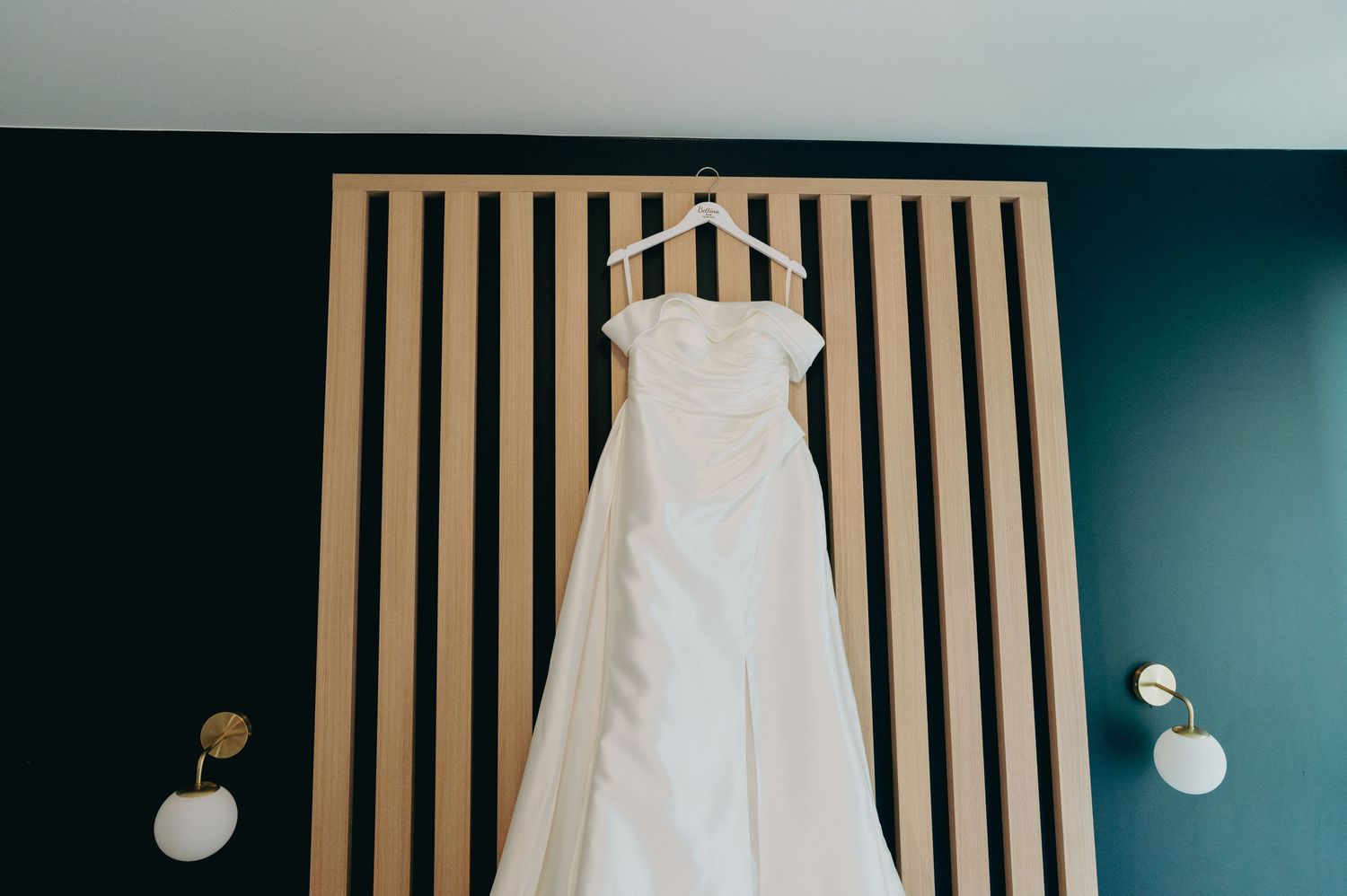 White wedding gown hanging on a wooden slat wall with gold sconce lighting in a modern room.
