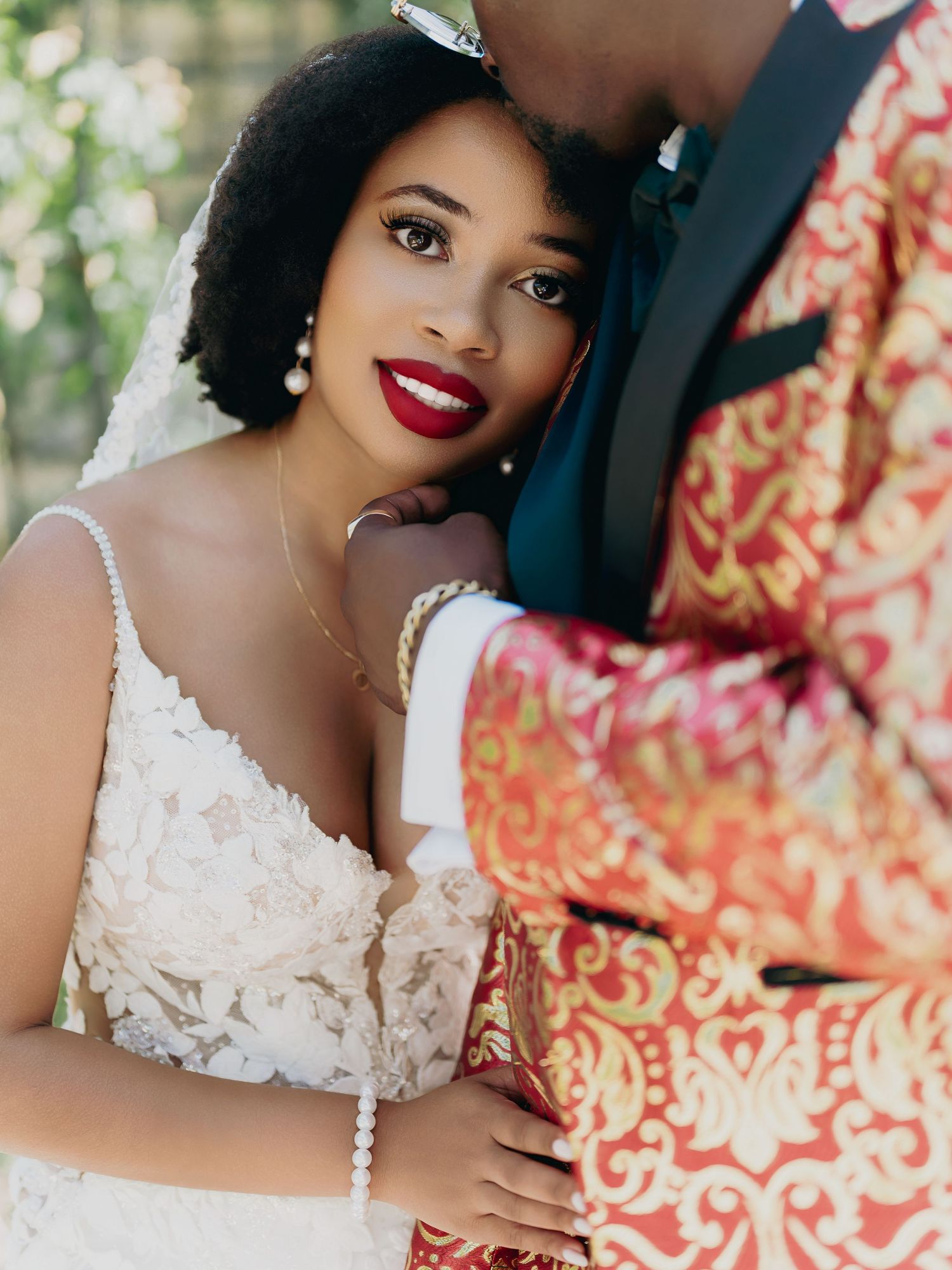 Close-up portrait of stunning bride in lace dress with red lips leaning against groom in ornate red blazer.