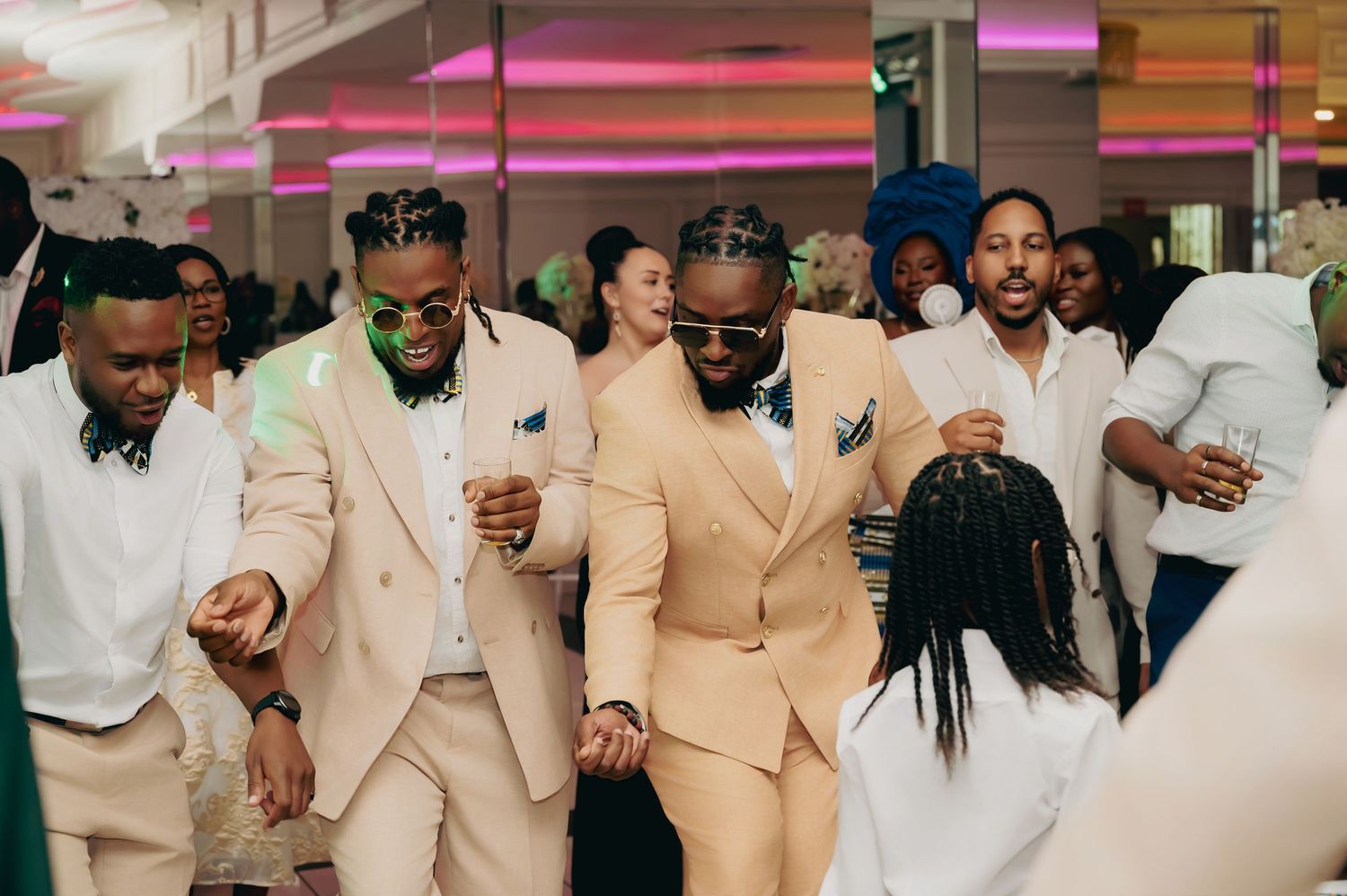 Groomsmen in beige suits dancing joyfully at a wedding reception with colorful lights.