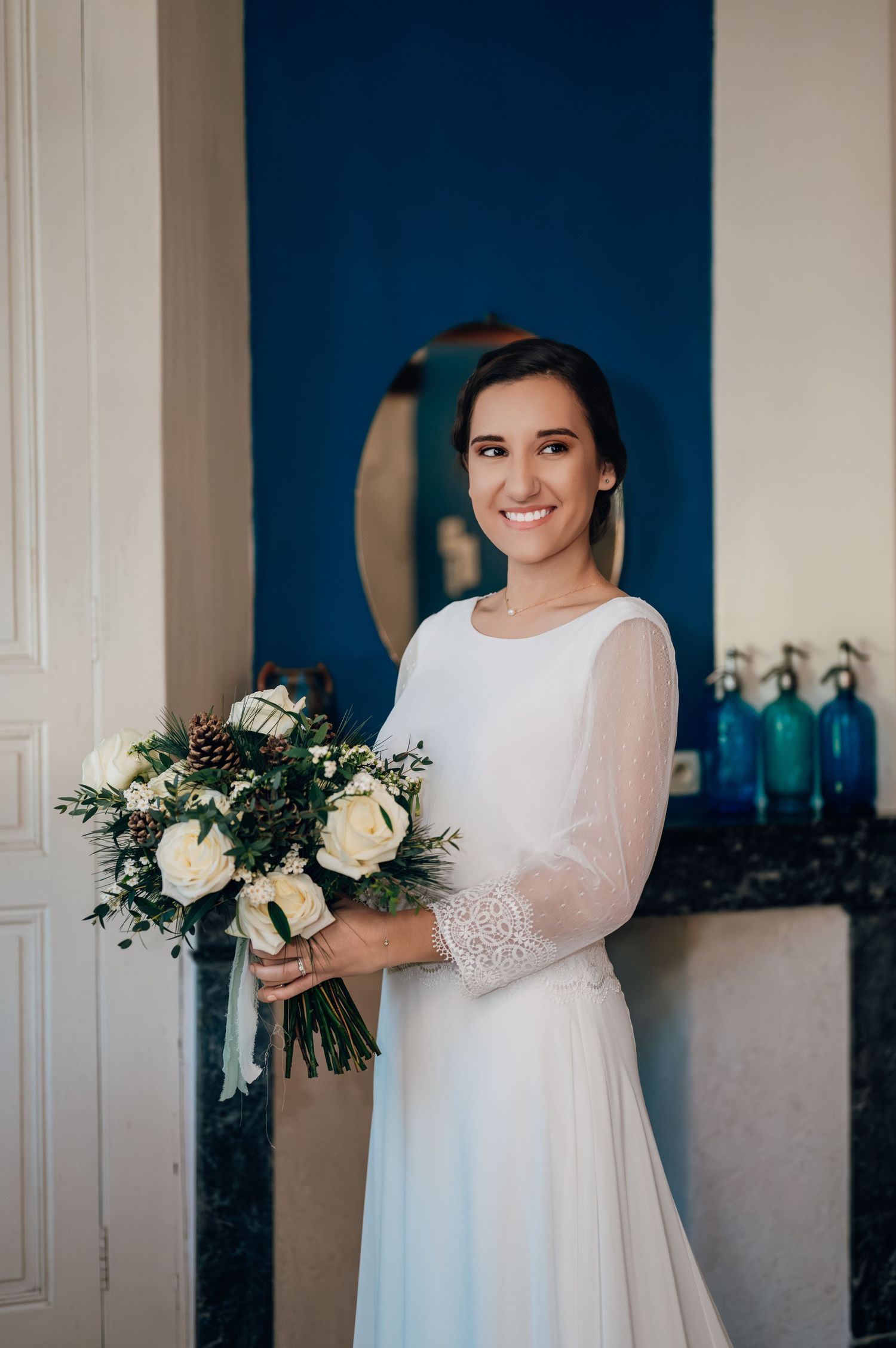 Smiling bride in white lace wedding gown holding a winter bouquet of white roses.