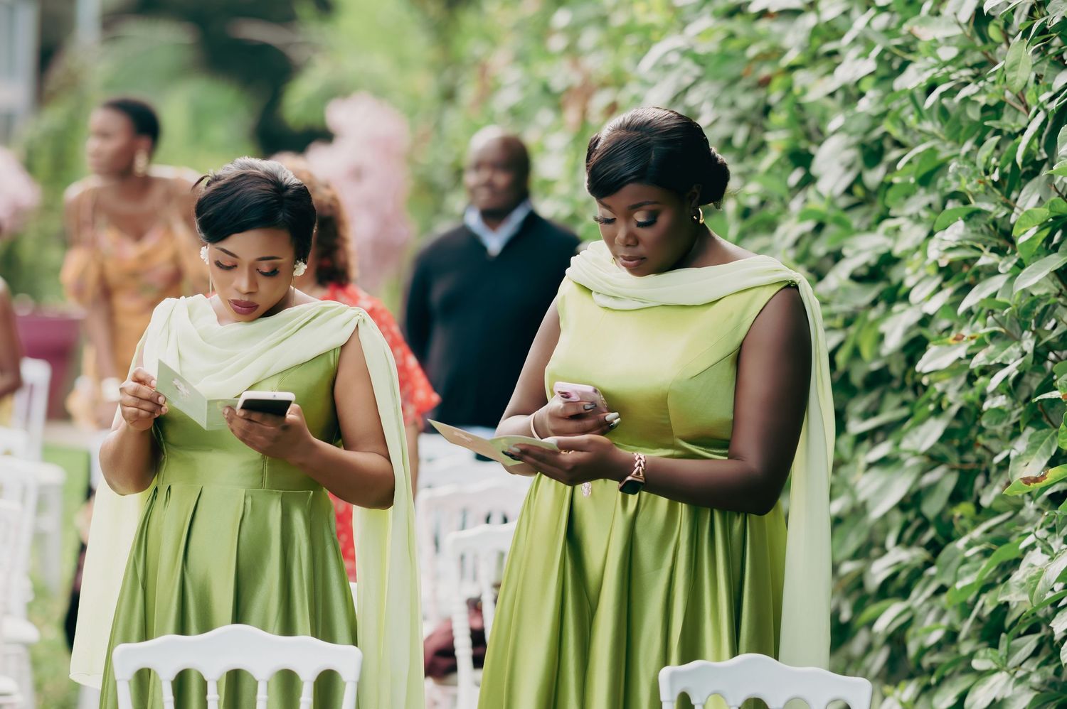Two bridesmaids in olive green dresses check their phones while standing beside white chairs at an outdoor wedding.
