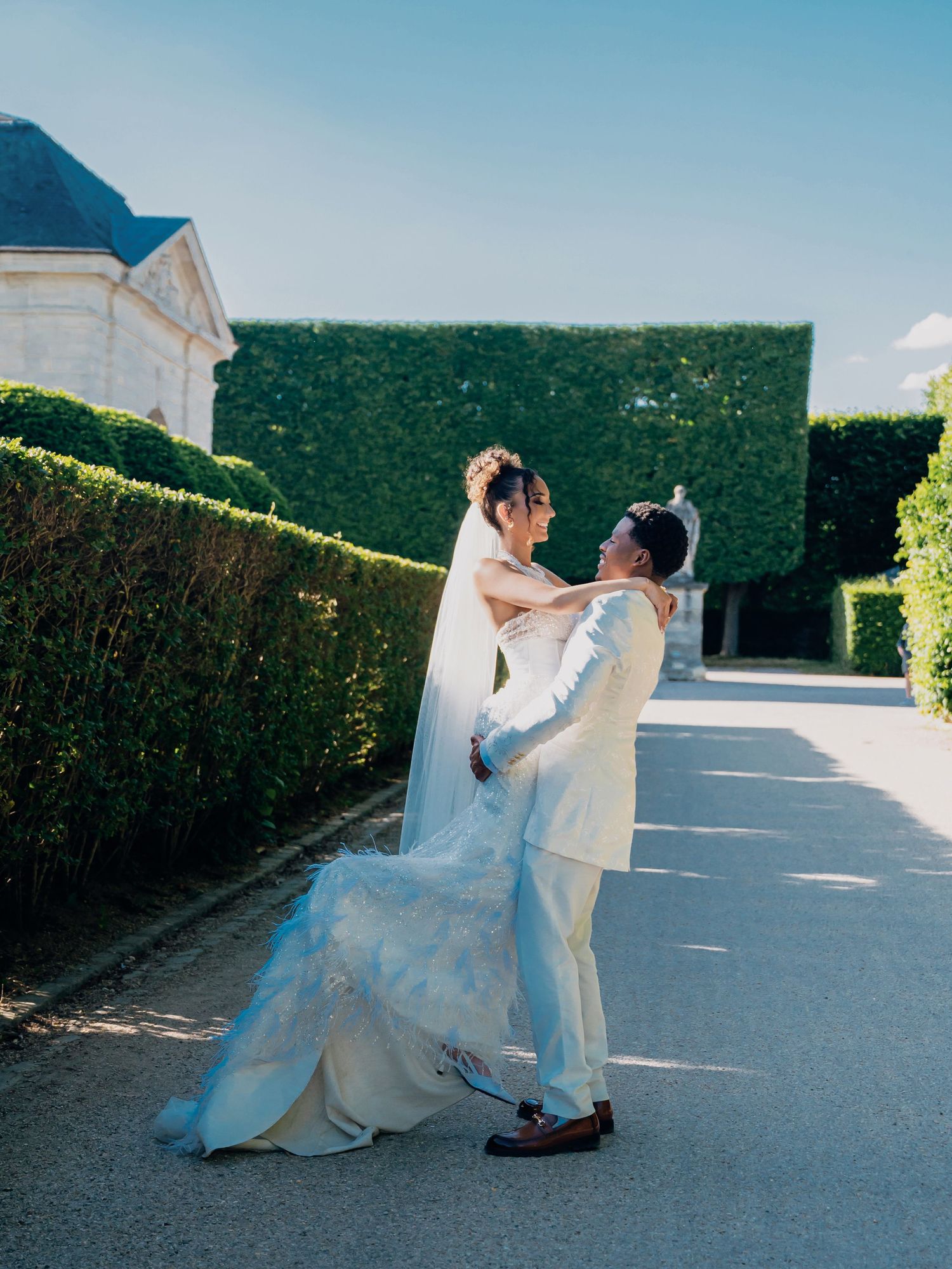 Groom lifting bride in white gown and veil outside French chateau with tall green hedges.