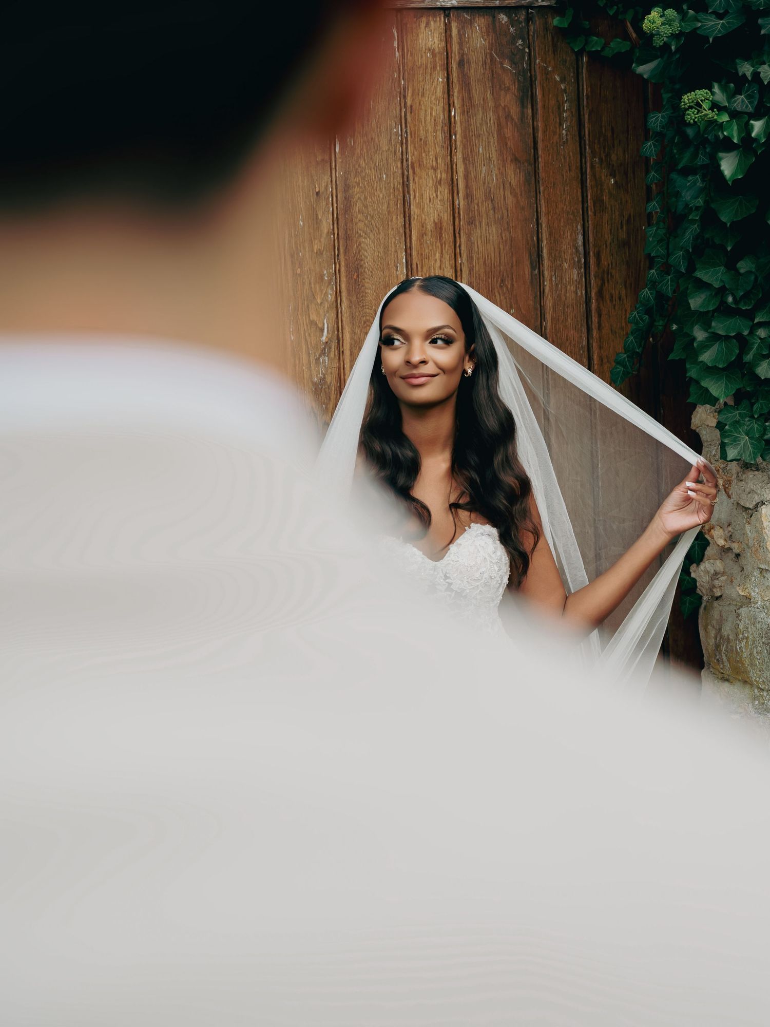 Beautiful bride in lace gown holding veil near rustic wooden door with green ivy backdrop.