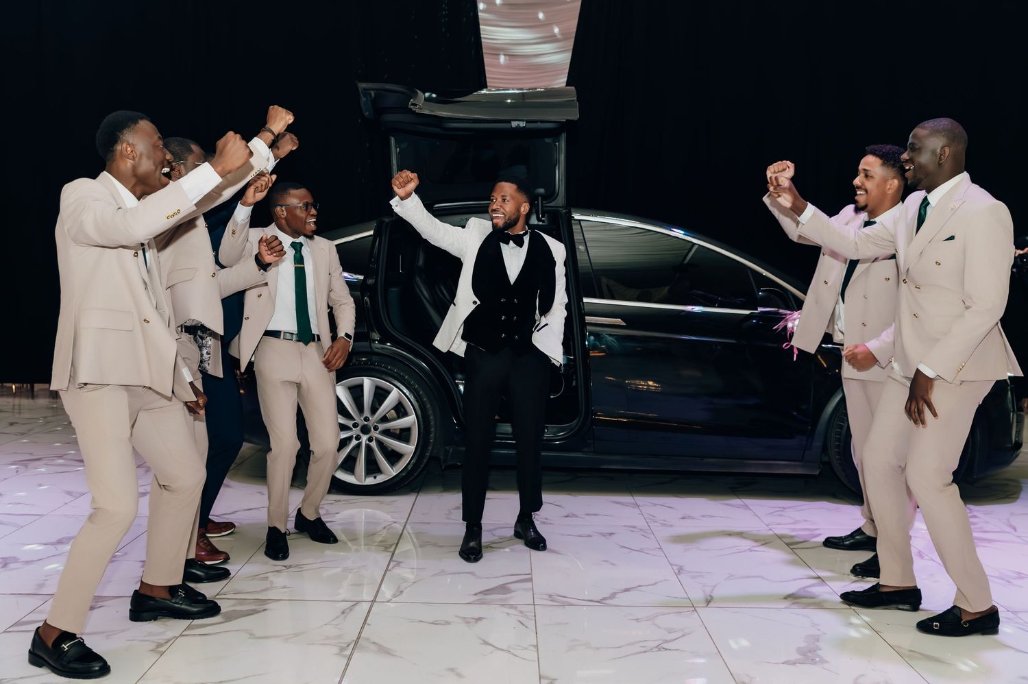 Groom in black tuxedo celebrates with groomsmen in cream suits beside a black Tesla SUV at a wedding.