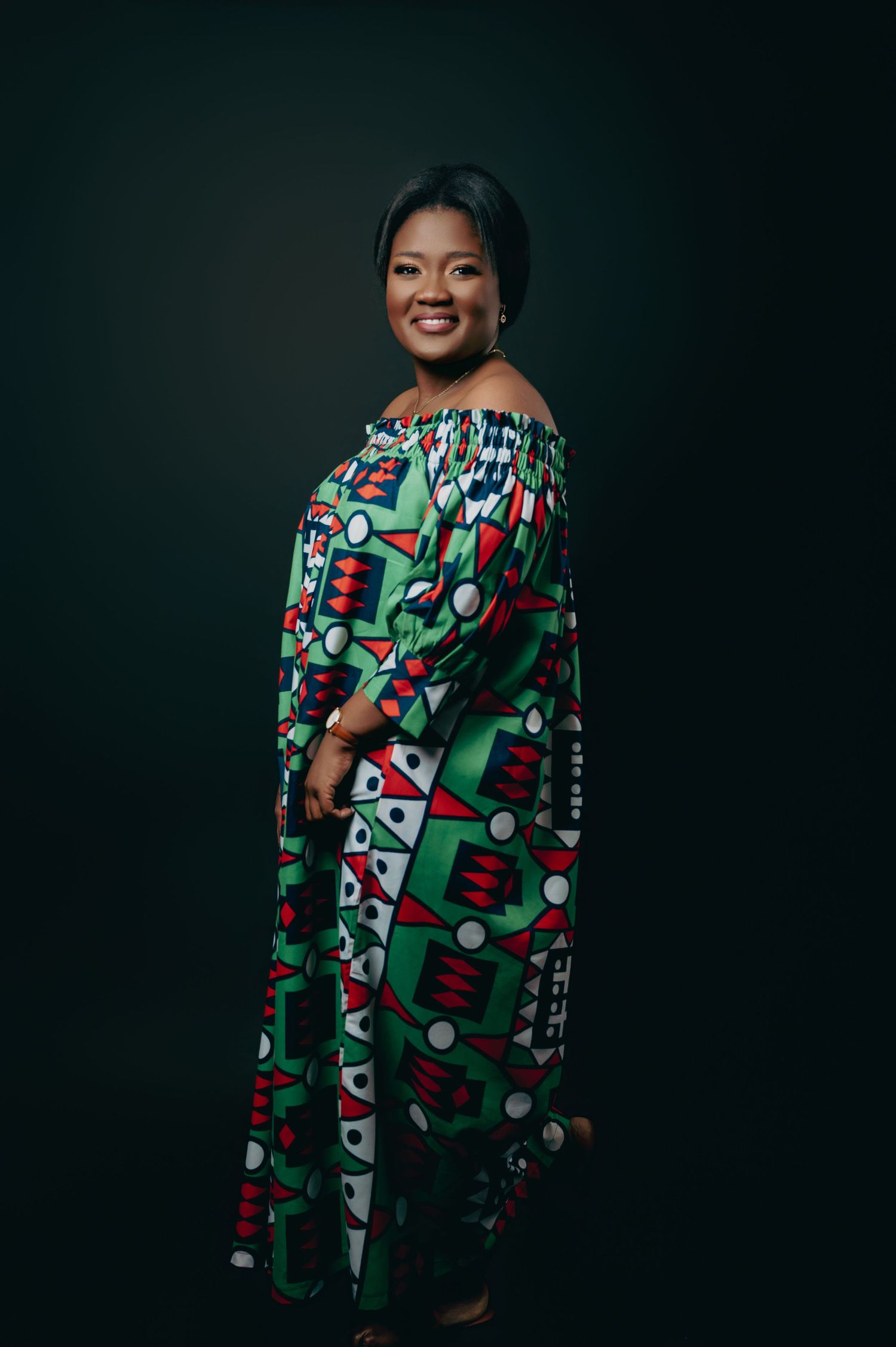 Elegant Black woman posing in vibrant green African print off-shoulder maxi dress against dark studio background.