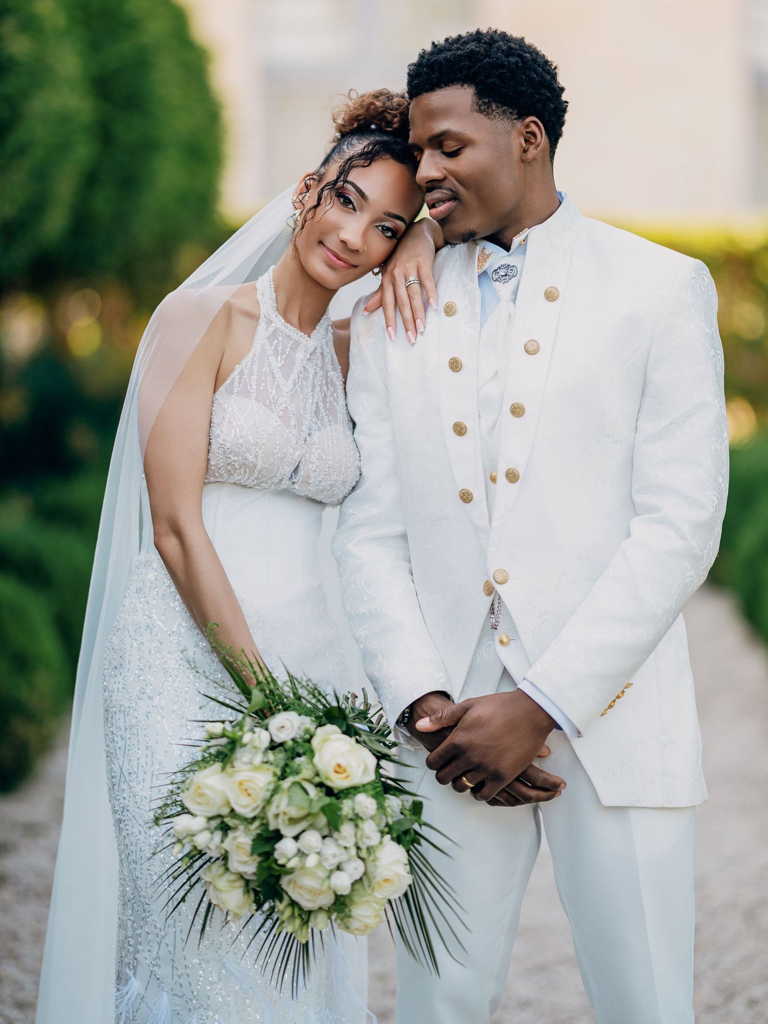 Couple poses in formal garden, bride in beaded halter gown with veil and groom in white double-breasted suit.