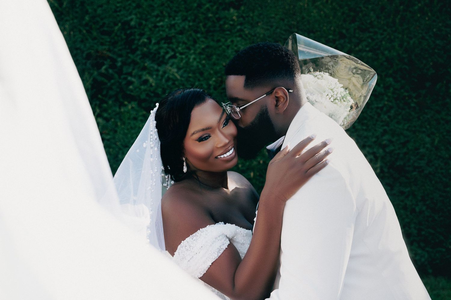 Romantic bride and groom nearly kissing outdoors surrounded by lush green hedge backdrop.