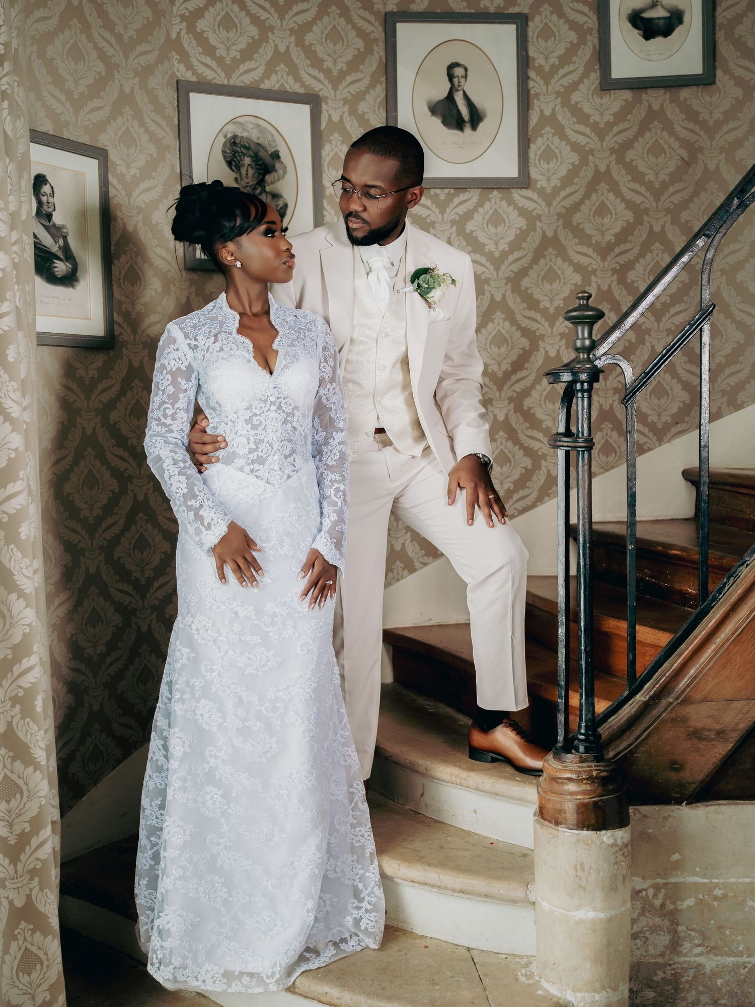 Elegant wedding couple posing on ornate staircase, bride in lace long-sleeve gown and groom in cream suit.