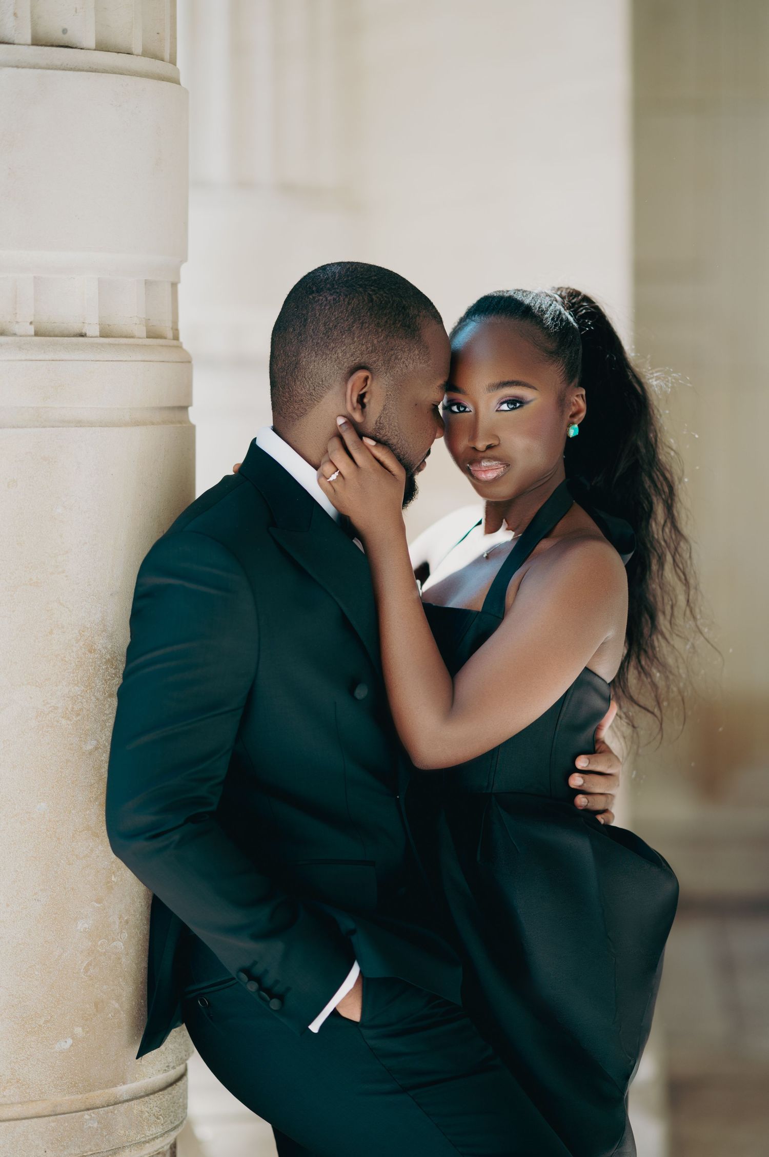 Black woman in black gown gazes at camera while embracing a man in a dark suit by a stone pillar.