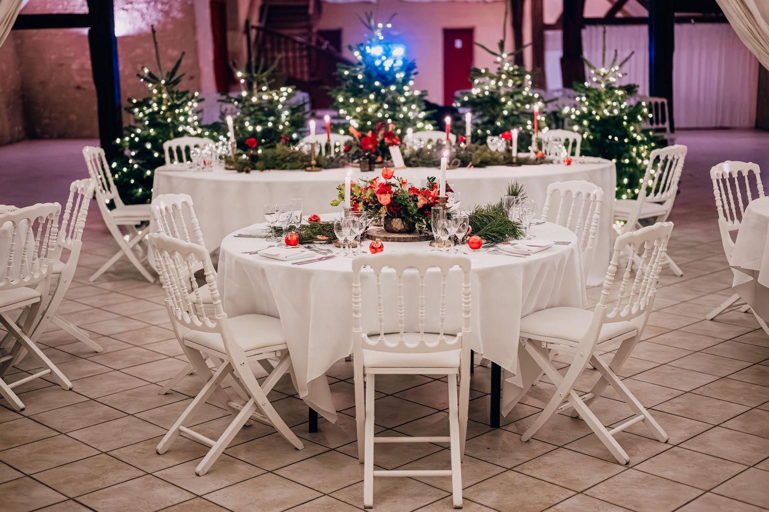 Christmas wedding reception table with white linens, red roses, candles, and decorated Christmas trees in a warmly lit venue.