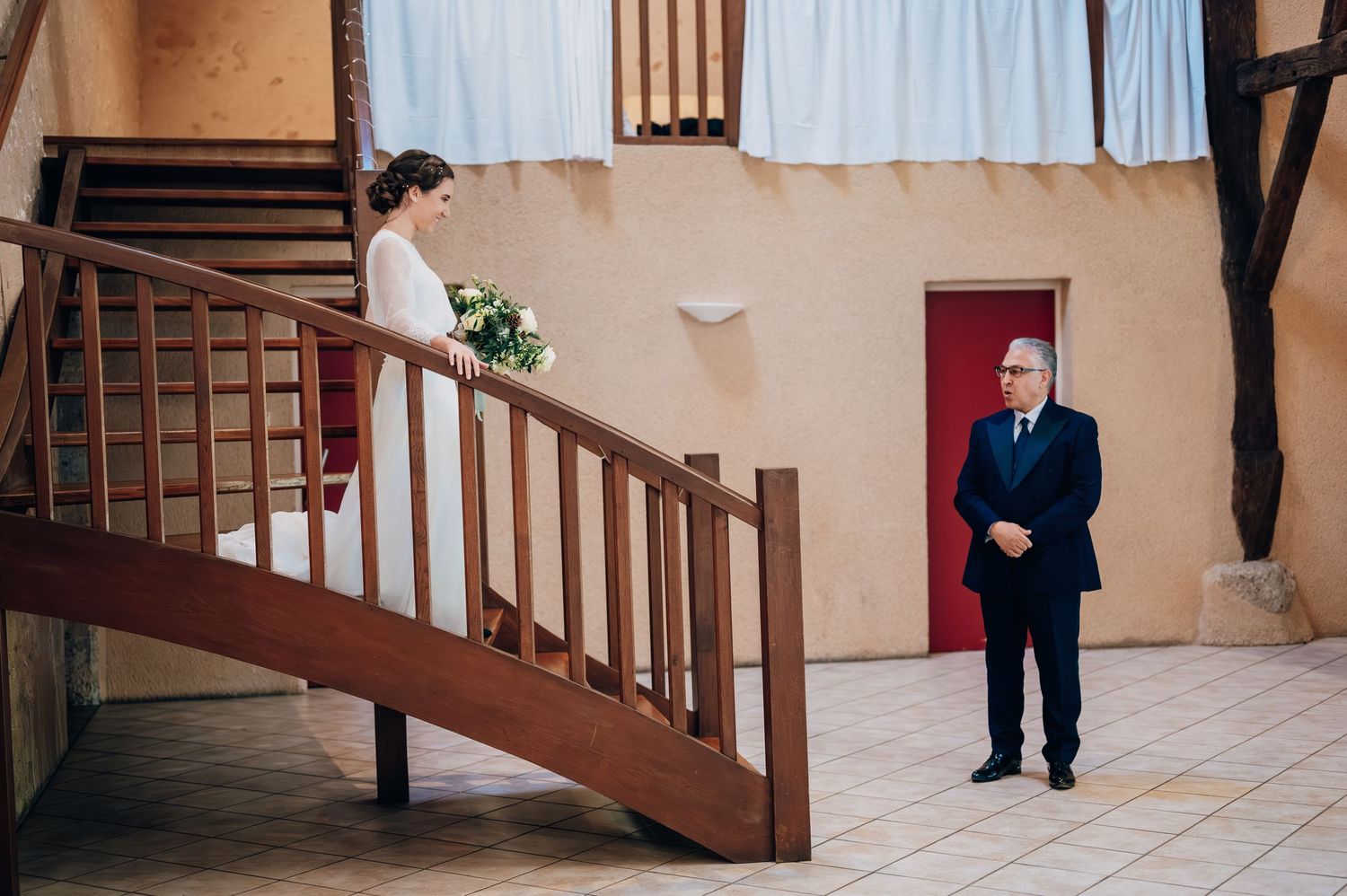 Bride in elegant white gown holding bouquet descends wooden staircase as a man in navy suit watches from below.
