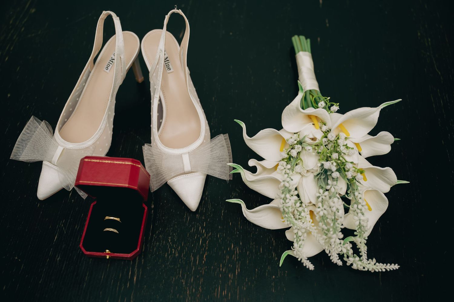 White bridal heels, red ring box with wedding bands, and a white calla lily bouquet on a dark surface.