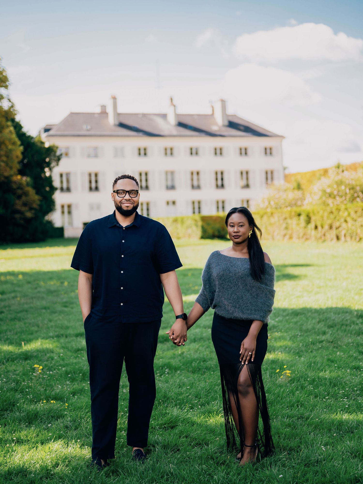 Engaged couple holds hands on green lawn with elegant white manor house behind them.