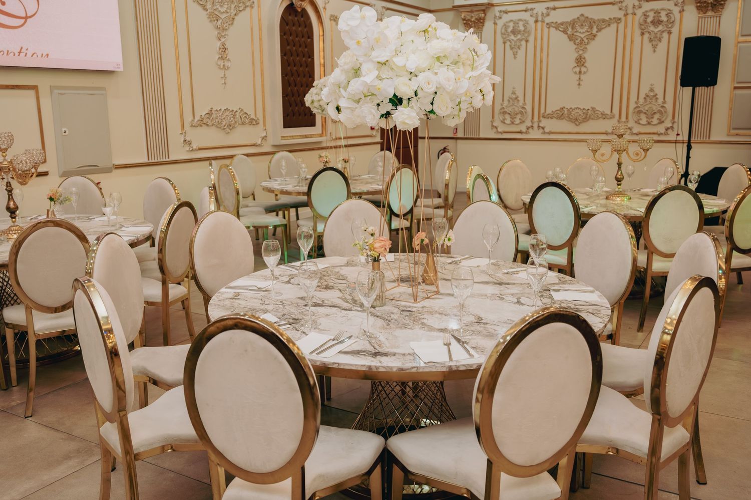 Elegant wedding reception table with marble top, gold chairs, and tall white floral centerpiece.