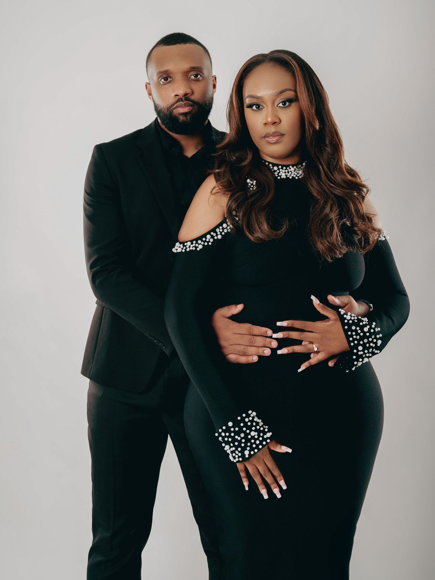 Pregnant woman in black beaded gown poses with partner in black suit during maternity shoot.